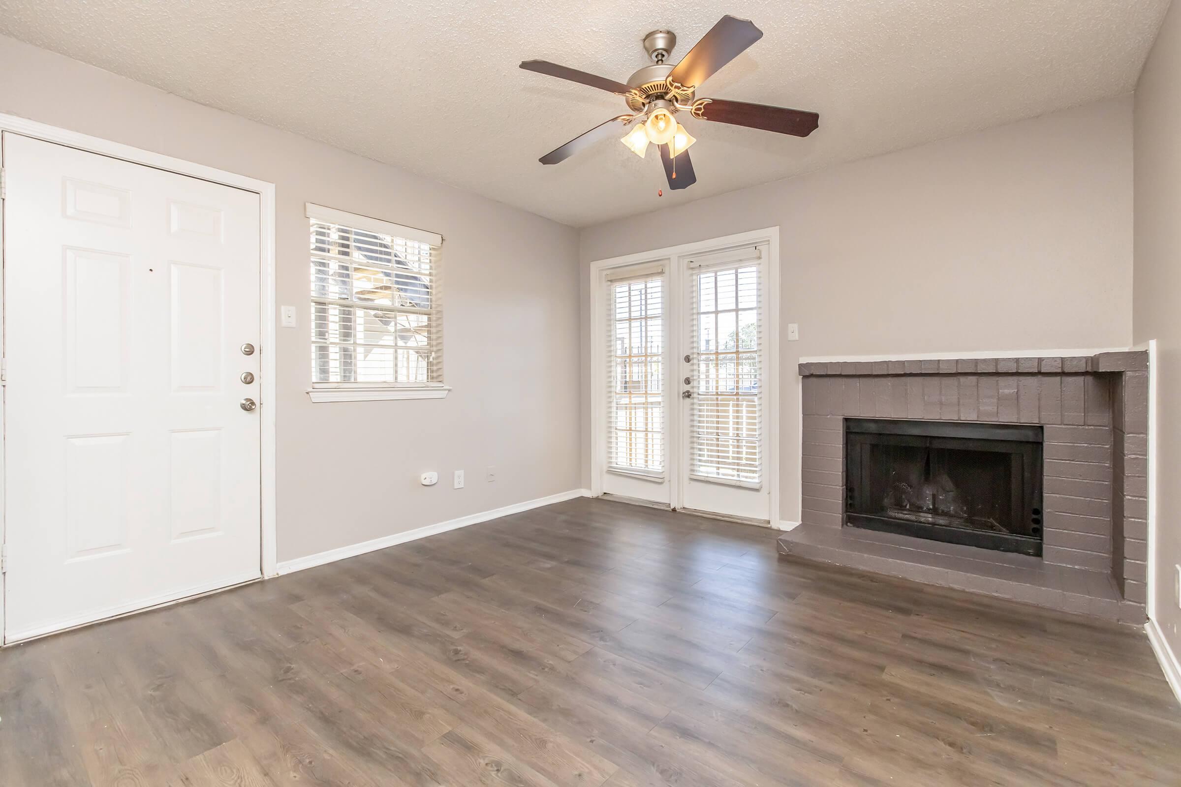 A spacious living room featuring a wood floor, a ceiling fan, and a fireplace. There is a front door to the left and two large windows on the right, providing natural light. French doors lead to an outdoor area, adding to the room's brightness and openness. The walls are painted in a light, neutral color.