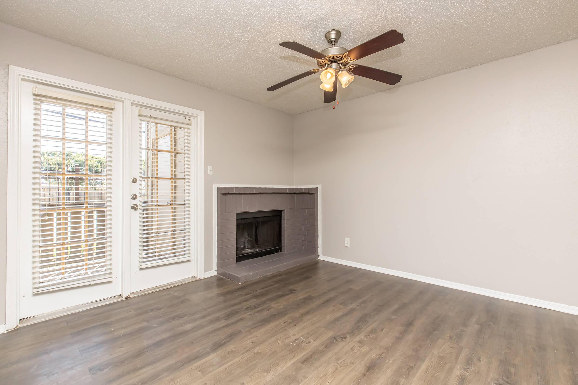 A spacious, empty living room featuring a ceiling fan, a fireplace with a gray surround, and large windows with white blinds. Light-colored walls and a modern wood-style floor enhance the bright and airy atmosphere. The room is ready for furniture and personalization.