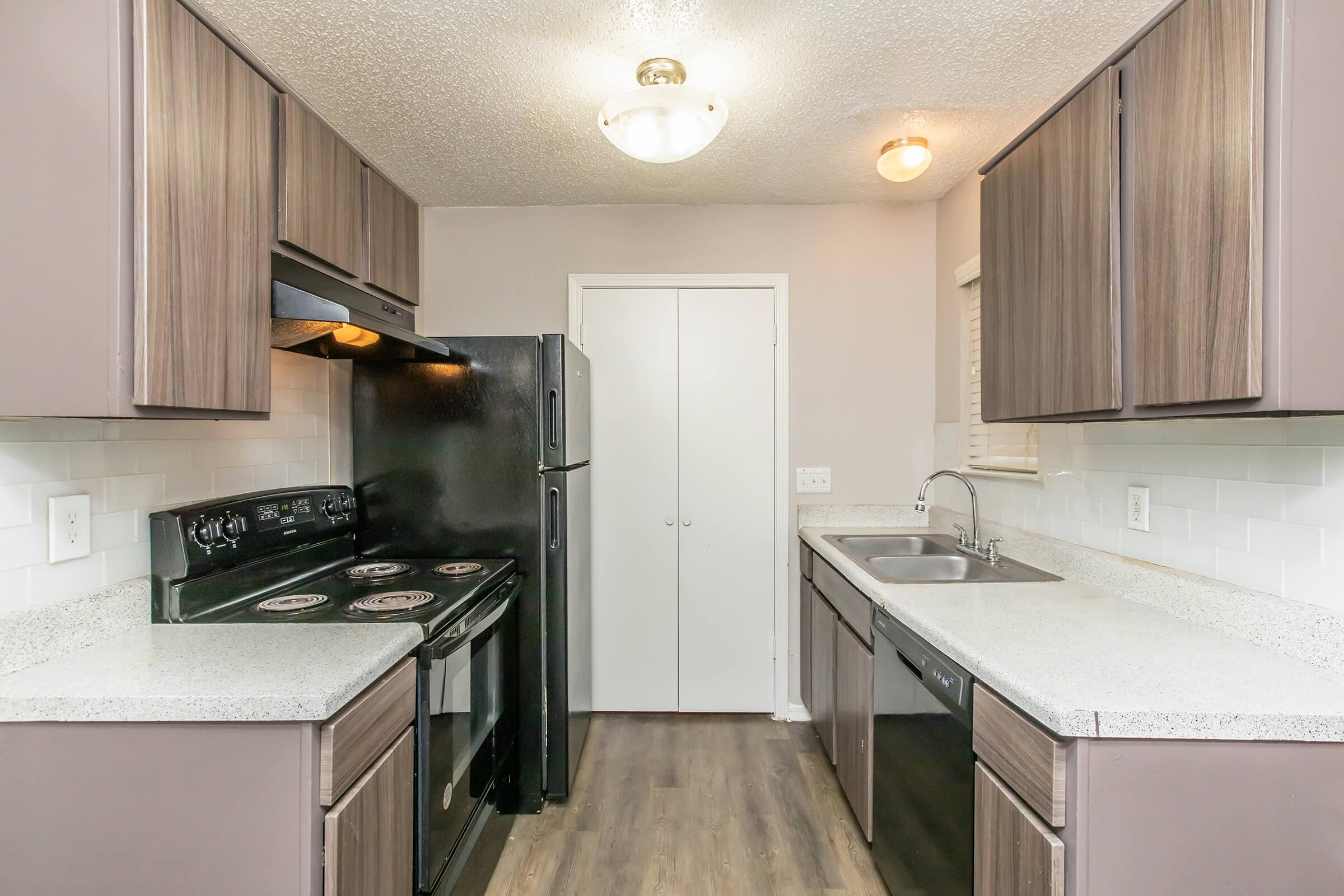 A modern kitchen featuring dark wood cabinets, a black stove and refrigerator, and a gray countertop. The space includes a stainless steel sink and a light-colored backsplash, with a doorway leading to another room. Soft lighting overhead enhances the contemporary feel of the kitchen.