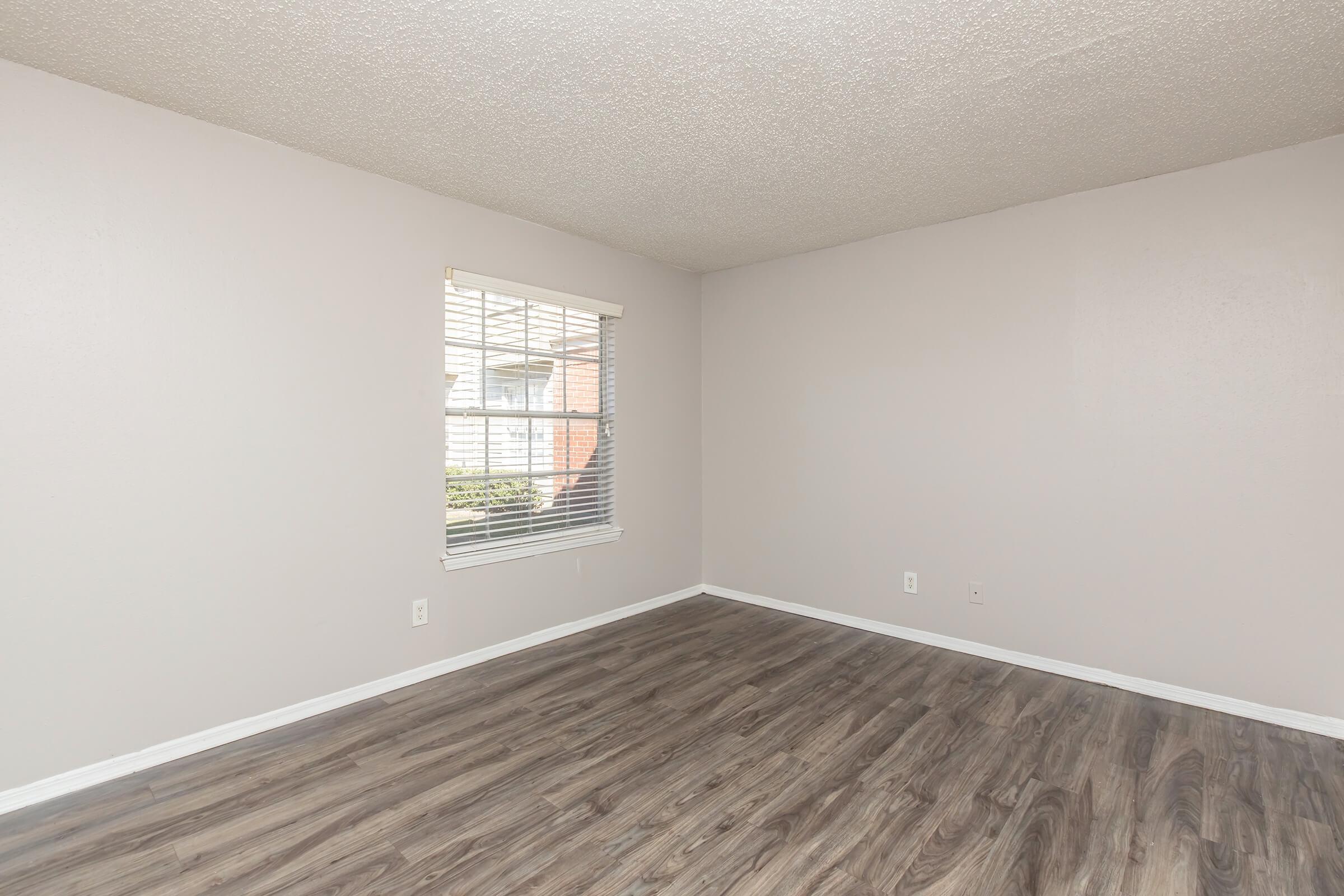 A spacious, empty room with light gray walls and a brown wood-like floor. A window with blinds allows natural light to enter. The room is uncluttered, providing a clean and modern feel, ideal for personal decoration or furnishing.