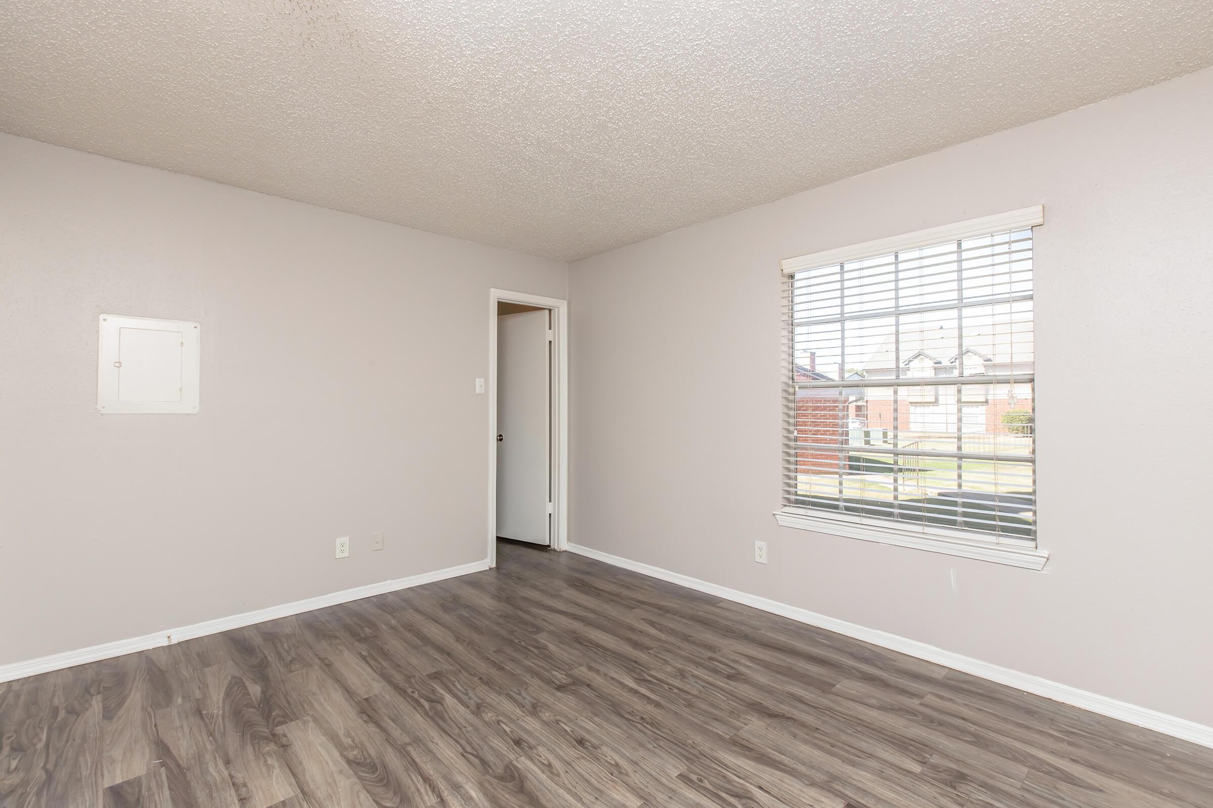 A well-lit empty room with light gray walls and a wood-like floor. A window with blinds allows natural light, and there's a white door leading to another space. A small wall panel is visible on the left. The overall aesthetic is clean and minimalistic.