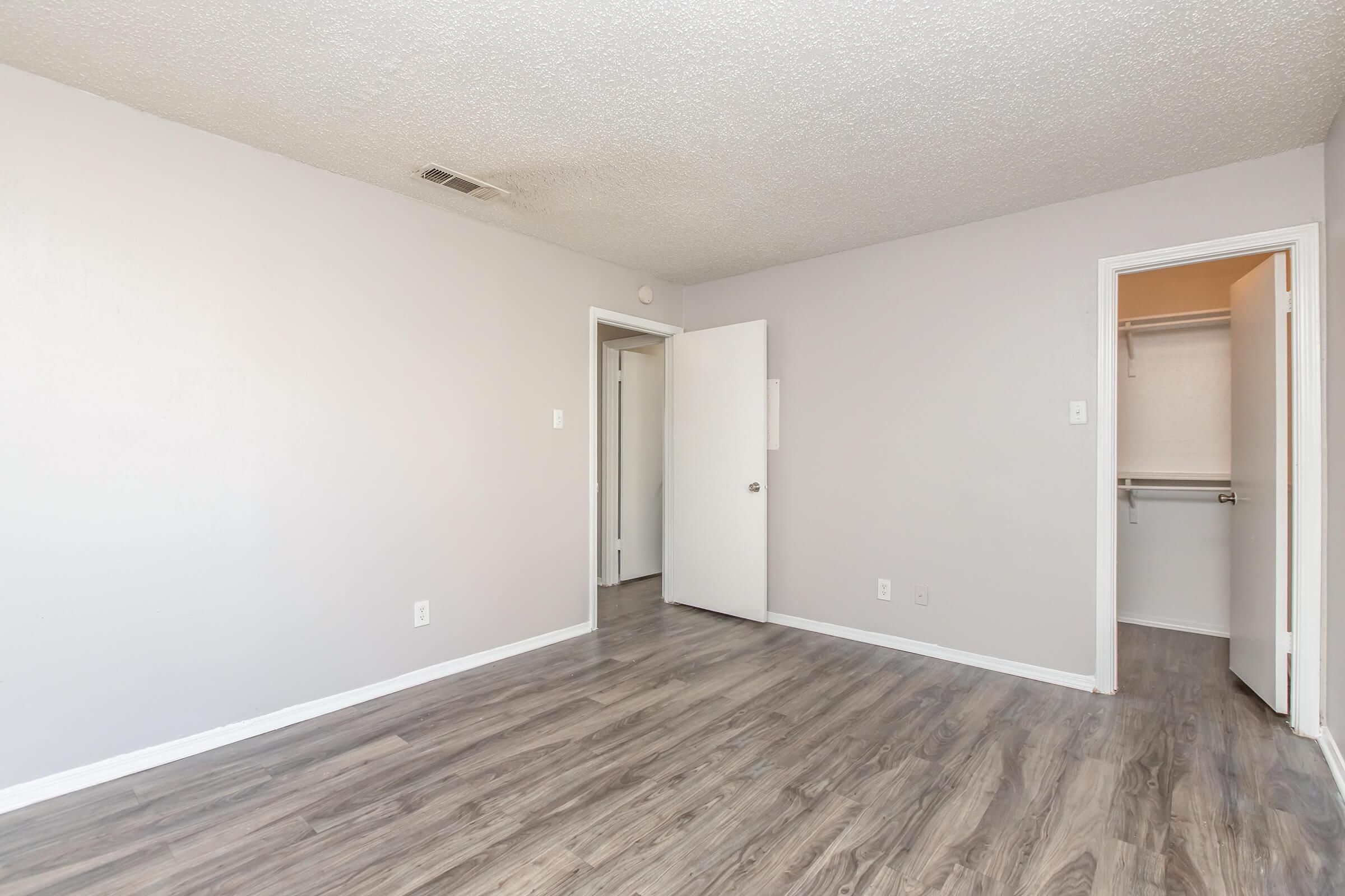 A spacious, empty room with light gray walls and wooden flooring. The room features two doors: one leading to another area and the other to a closet. Natural light is visible, creating a bright and inviting atmosphere. The ceiling has a textured finish, enhancing the room's aesthetic appeal.