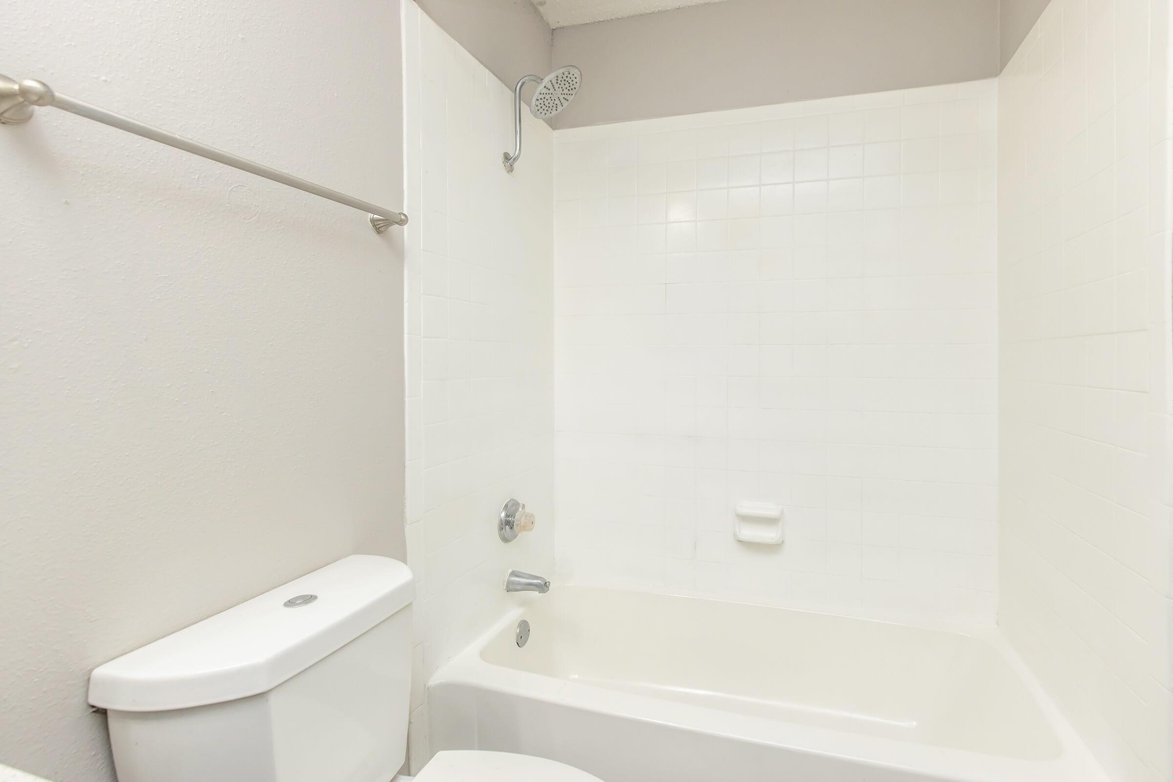 A clean, modern bathroom featuring a white bathtub with a showerhead, a simple wall-mounted soap dish, and a white toilet. The walls are painted in a light color, and the tiled area is bright and well-maintained.
