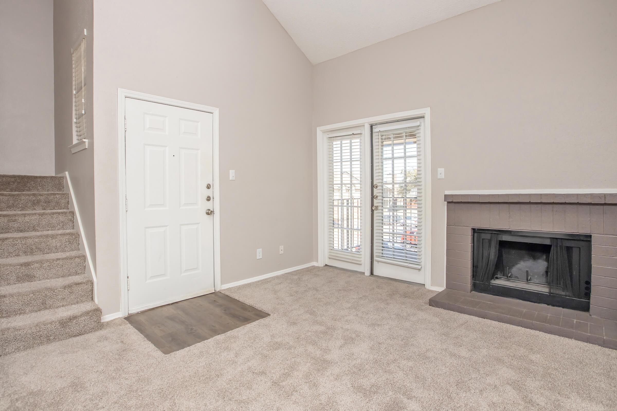 Spacious living area with light beige walls and carpet, featuring a fireplace with a brick surround. There is a door leading to an outdoor space and a staircase on the left. Natural light enters the room through sliding glass doors adorned with white blinds.