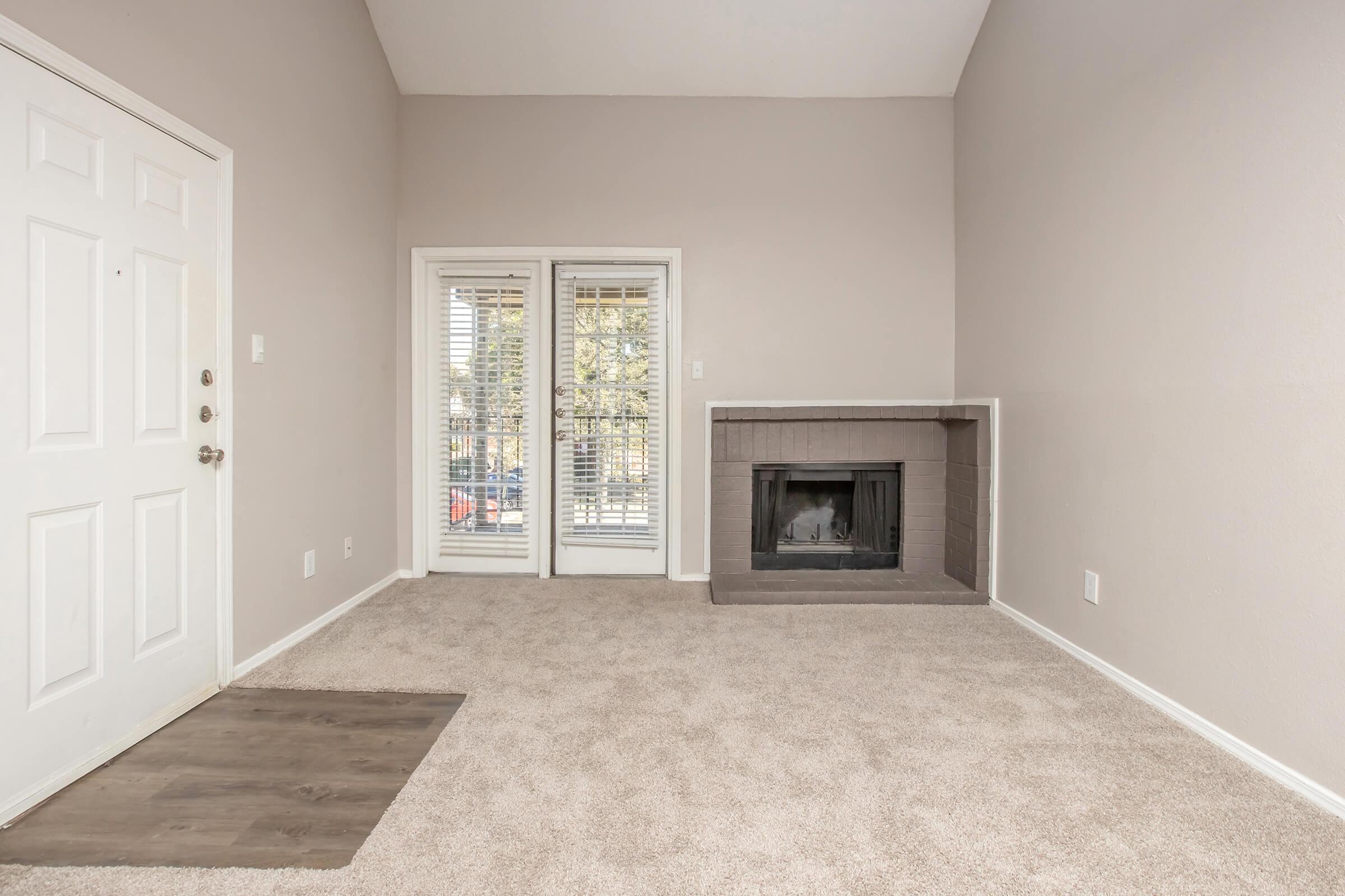 A spacious, light-filled room featuring a beige carpet, a white front door, and large windows with blinds. There's a cozy fireplace on one side and a small entryway with a different flooring material. The walls are painted in a soft gray color, creating a neutral and welcoming atmosphere.