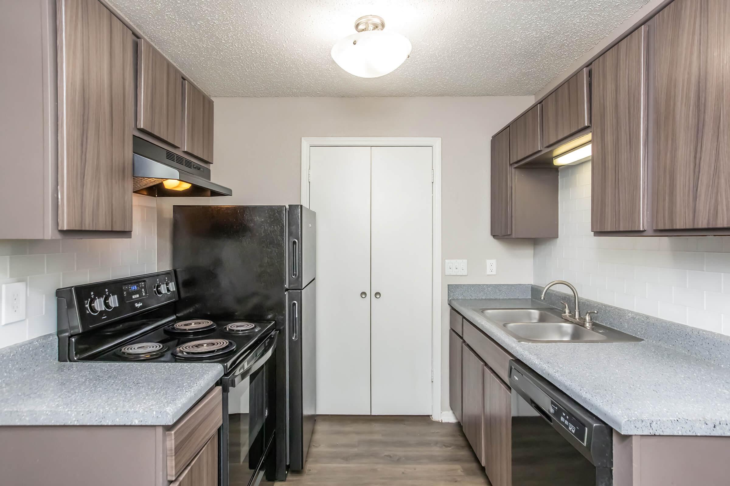 A modern kitchen featuring dark wooden cabinets, a black stove and refrigerator, and a double sink. The walls are light-colored, and the countertops are gray. There is a white door leading to another space, and the kitchen is well-lit by a ceiling fixture. The overall design is functional and contemporary.
