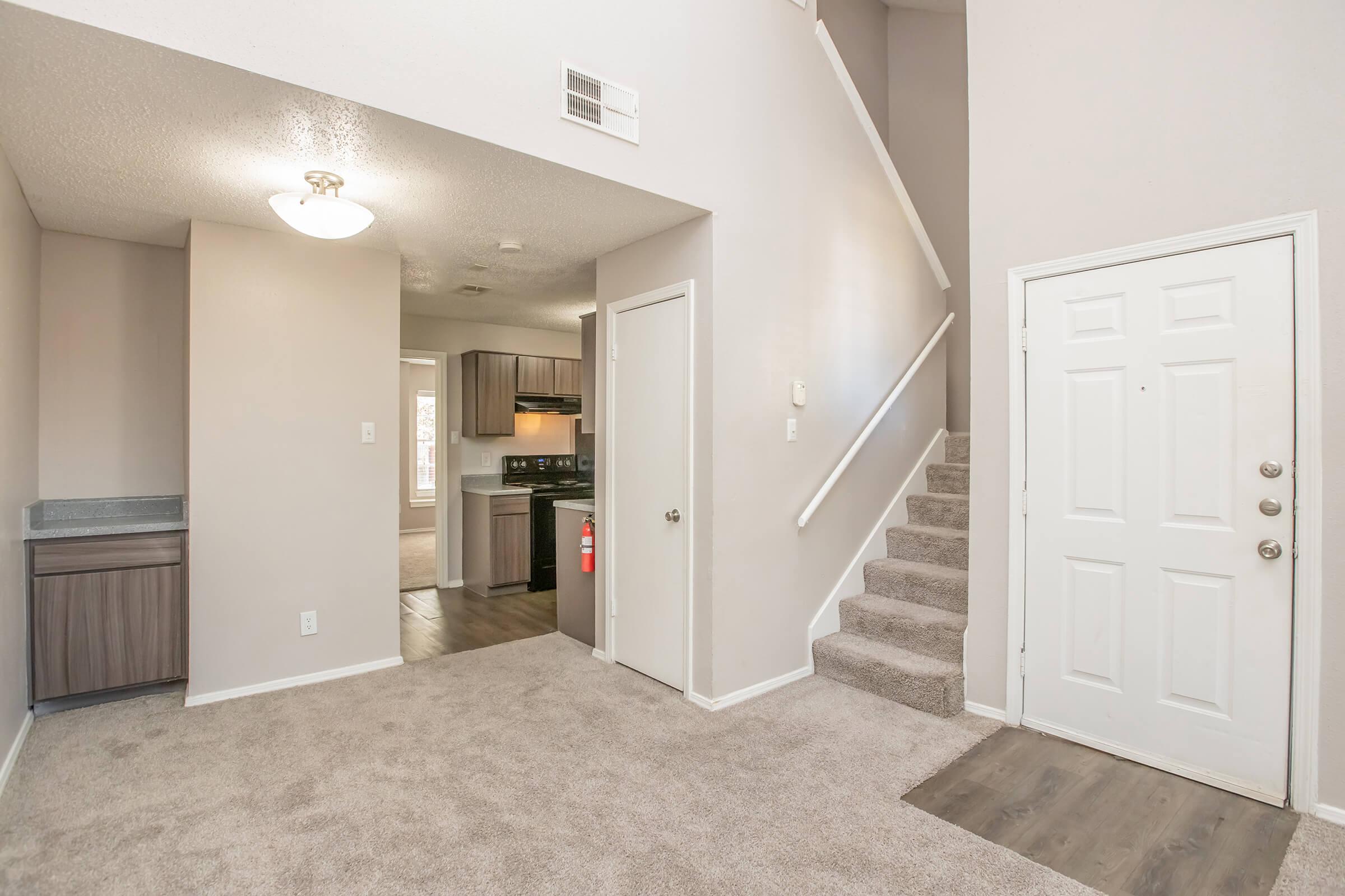 A view of an inviting entryway in a home, featuring beige carpet, a staircase leading up, and a doorway leading to a kitchen area in the background. Natural light enters from a window, and there’s a small counter to the left. The front door is white, with a simple design and a welcome mat.
