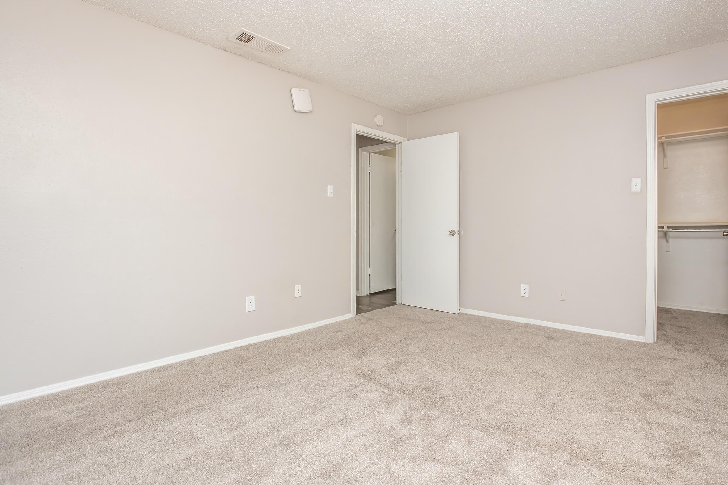 Empty room with light gray walls and tan carpet. A door is slightly ajar, leading to a closet on the right. The space has minimal decor, creating a clean and open feel. Small wall-mounted speaker or light fixture visible on the left side. Natural light may be entering from an unseen window.