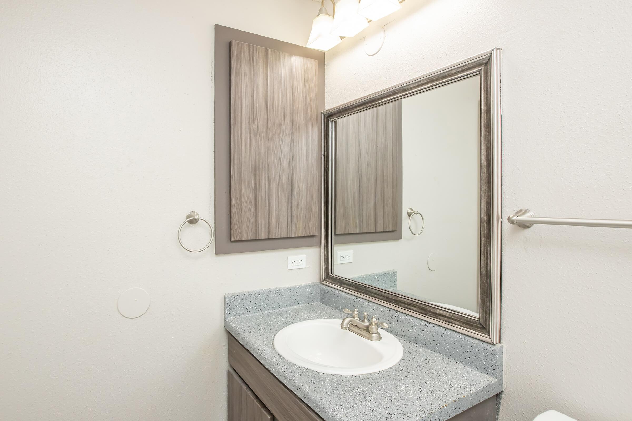 A modern bathroom featuring a gray countertop with a round sink, a large mirror above, and a decorative light fixture. There is a wooden cabinet above the sink, a towel ring on the wall, and a clean, minimalist design with light-colored walls.