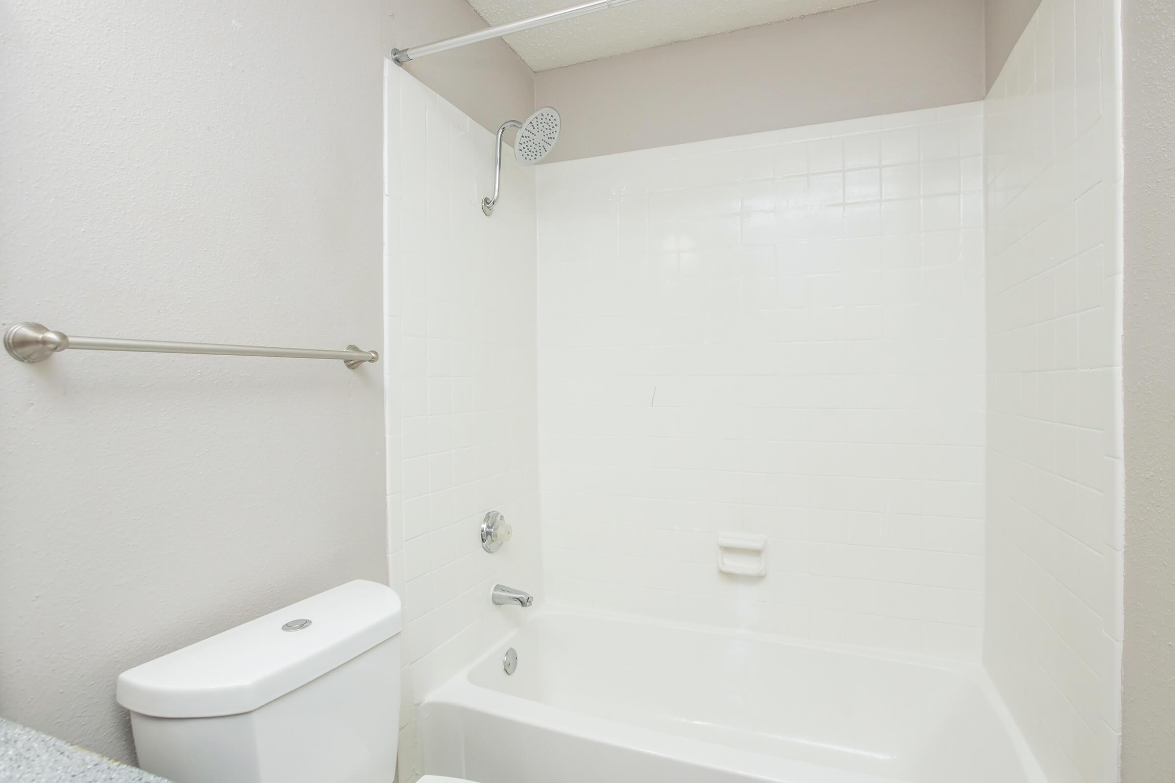 A clean and simple bathroom featuring a white bathtub with a showerhead, a wall-mounted grab bar, and a toilet. The walls are a light color, and the space is well-lit, creating a bright and airy atmosphere.
