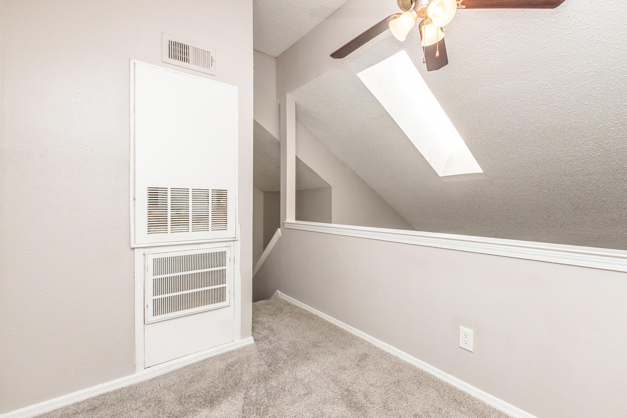 A small, well-lit room featuring beige walls and carpet. There is a ceiling fan with light fixtures and a skylight that provides natural light. A white wall vent is visible, and stairs lead to another level, creating an inviting atmosphere.