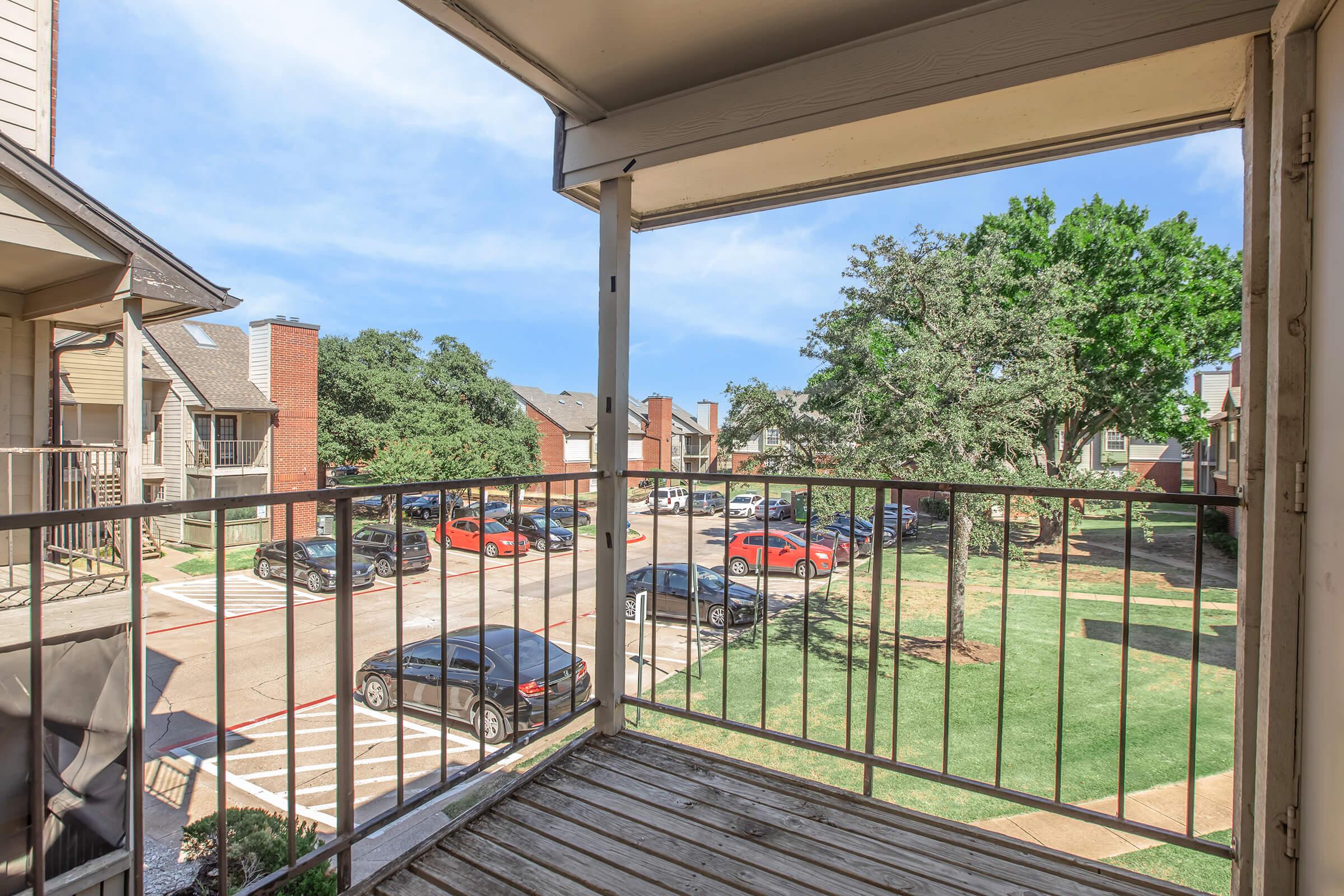 View from a balcony overlooking a parking lot surrounded by landscaped grounds. Vehicles parked in a row, with residential buildings in the background and lush green trees nearby. The sky is clear and blue, indicating a pleasant day.