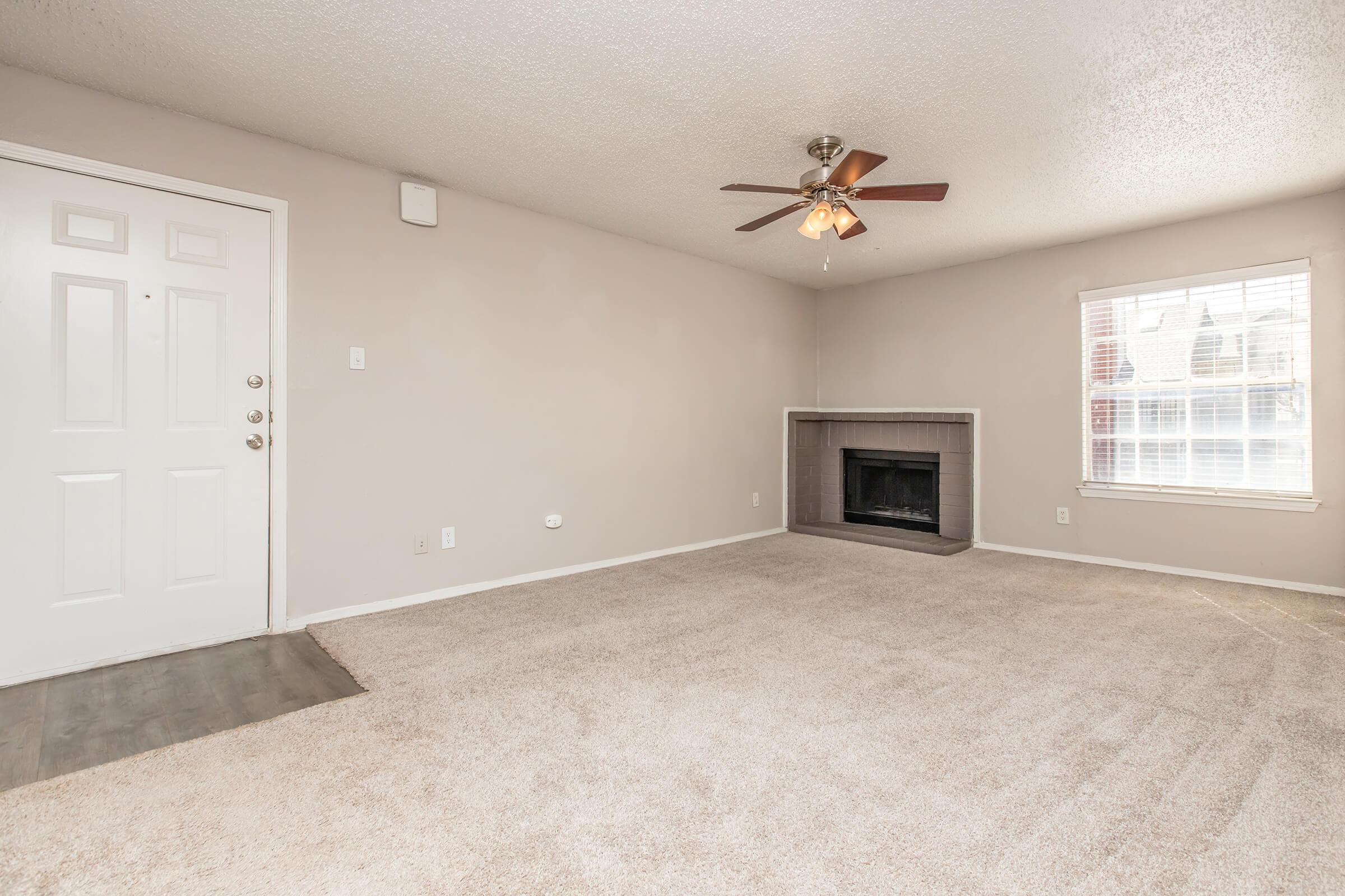 A spacious, empty living room featuring light-colored walls, a ceiling fan, and a decorative fireplace. The room has a large window allowing natural light and a doorway leading outside. The flooring includes carpet and a small section of wood. The overall atmosphere is bright and open.