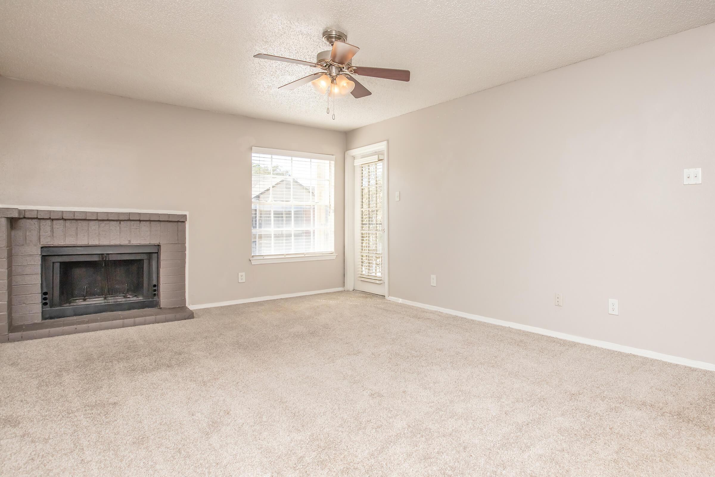 A spacious living room with light beige carpet, a ceiling fan, and a fireplace. The room features a large window letting in natural light, and there is a sliding glass door leading outside. The walls are painted in a neutral color, creating a calm and inviting atmosphere.