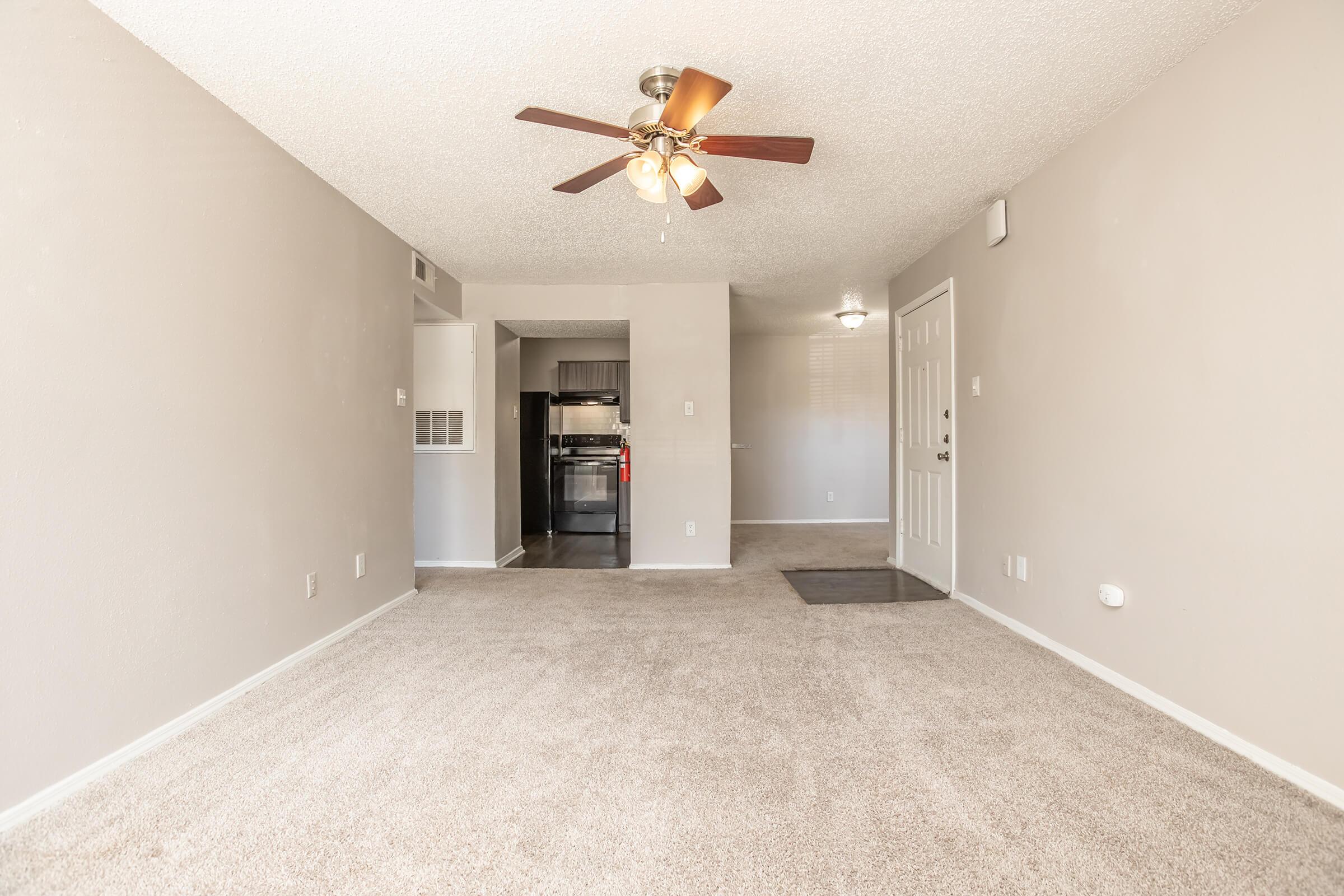 A spacious living area with light beige carpet, a ceiling fan, and textured walls. A dark kitchen is visible in the background, featuring black appliances. The entrance door is on the right, and there’s a small entrance mat on the floor. Natural light brightens the room, creating a welcoming atmosphere.
