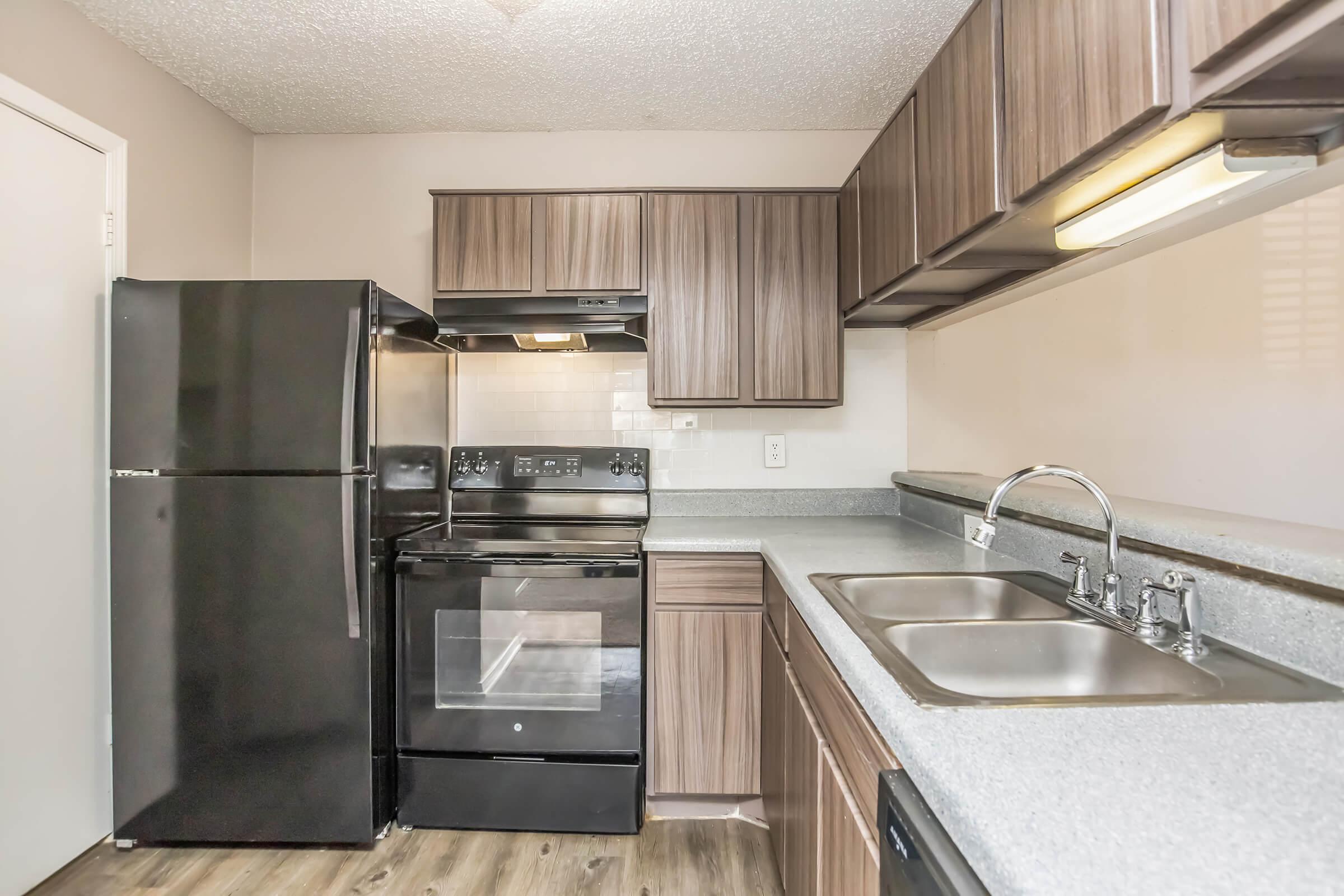 Compact kitchen featuring dark wood cabinets, a black refrigerator, and a black stove with a matching hood. The countertop is gray with a double sink, and there are white tiles on the backsplash. Natural light is coming through a small window, illuminating the space.