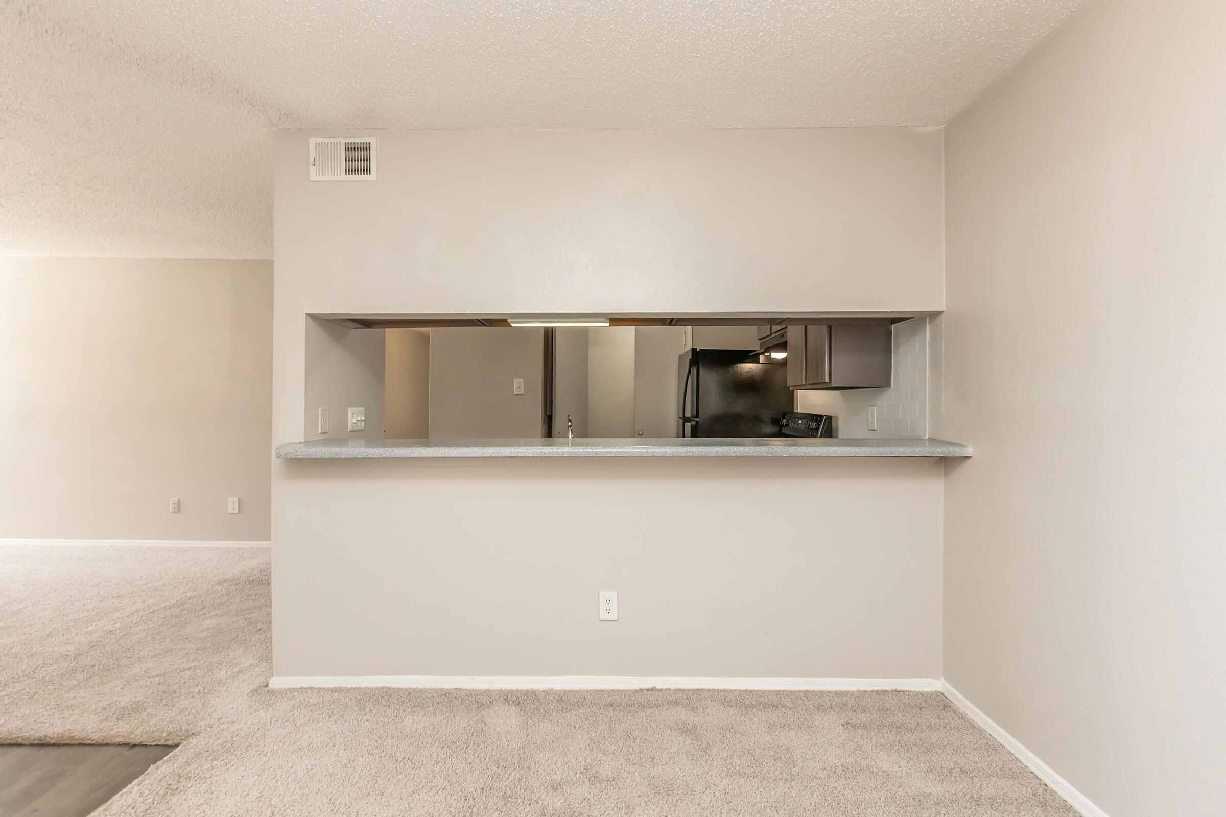 Interior view of a spacious living area featuring a light-colored carpet. In the foreground, there is an opening to a kitchen with gray countertops and modern appliances visible in the background. The walls are painted in neutral tones, enhancing the bright and airy atmosphere of the space.