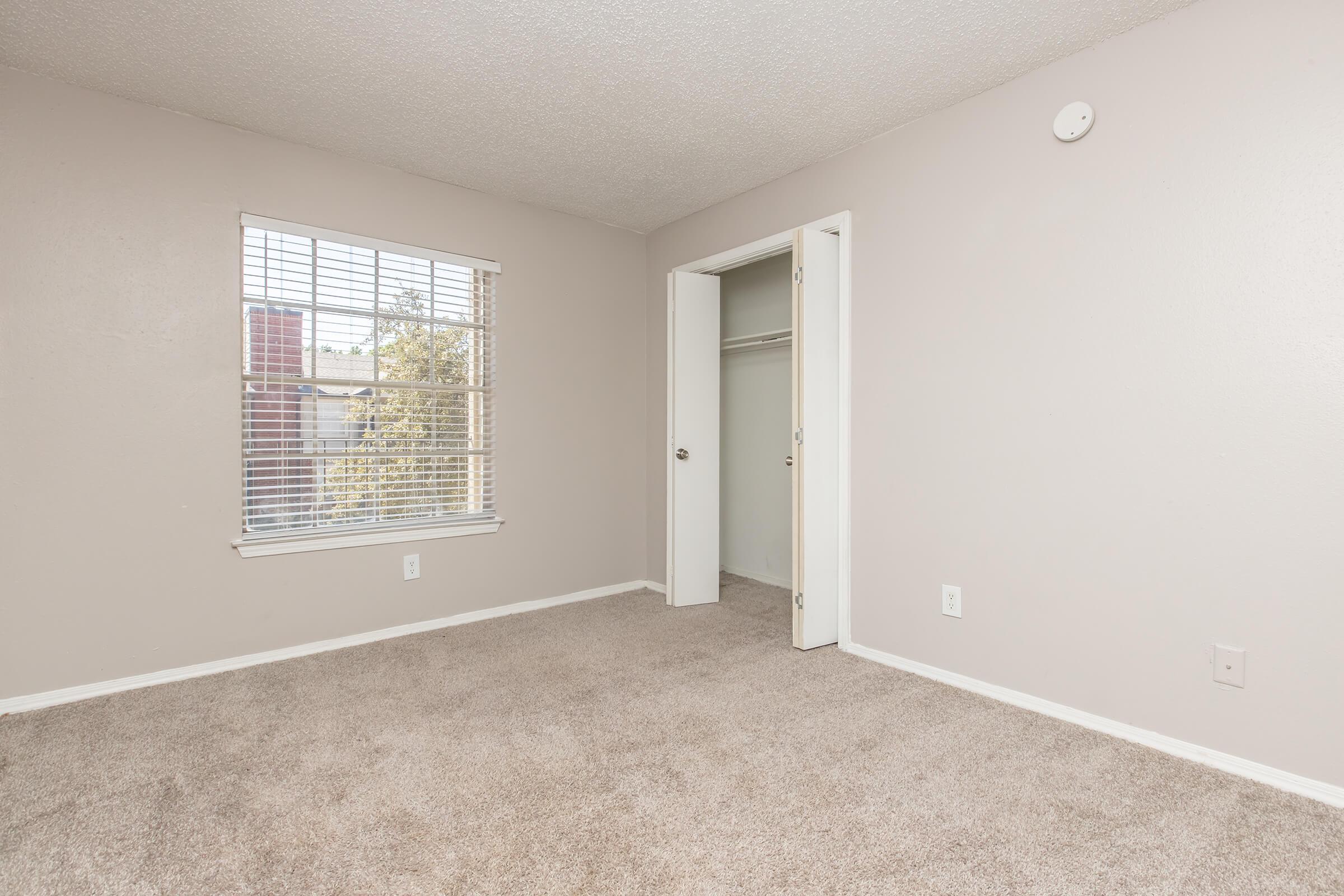 A clean, empty bedroom featuring light-colored walls, a window with blinds allowing natural light, and a closet with an open door. The room has beige carpet and a simple, minimalist design, creating a spacious feel.