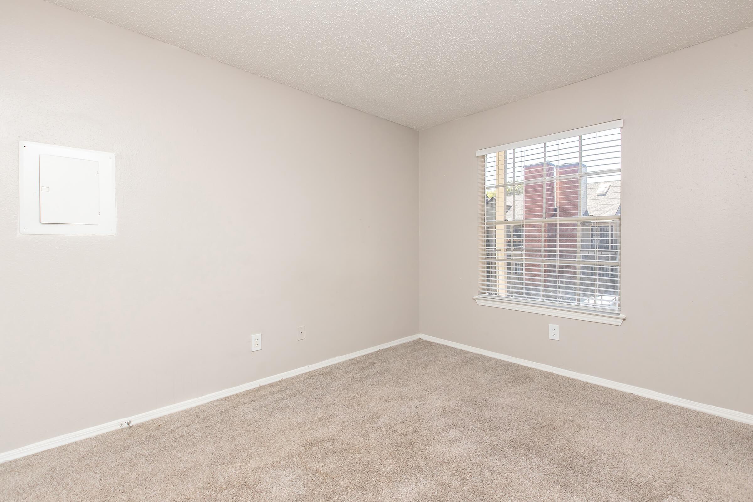 Empty room with light beige walls and carpet. A window with blinds is on the right side, allowing natural light to enter. The ceiling is textured, and there is a small electrical panel on the left wall. The space appears clean and ready for furnishings.
