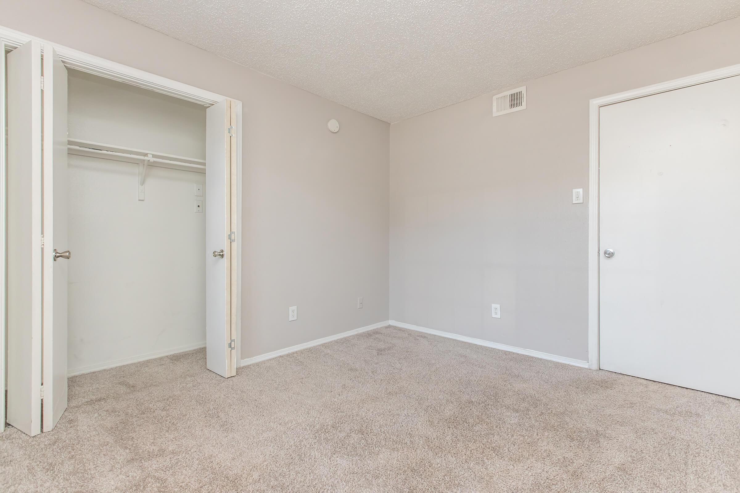 A spacious, empty bedroom featuring beige carpet, light gray walls, and two doors—one leading to a closet and the other a standard door. The room has no furniture, creating a minimalist and airy feel, with natural light illuminating the space.