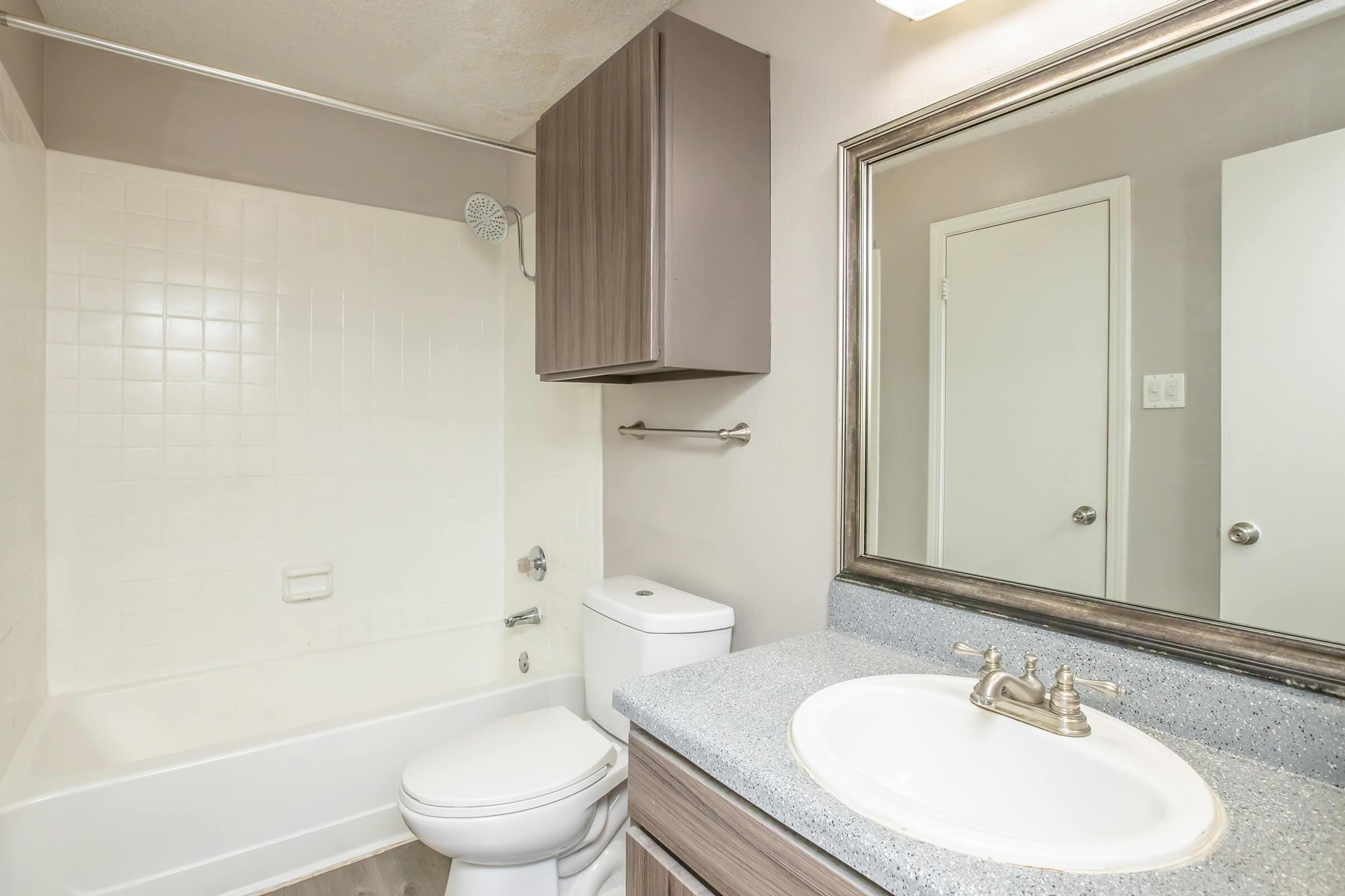 A clean and well-lit bathroom featuring a bathtub with a showerhead, a modern toilet, and a sink with a granite countertop. The walls are painted in a neutral color, and there is a large mirror above the sink. A cabinet is mounted above the toilet for storage.