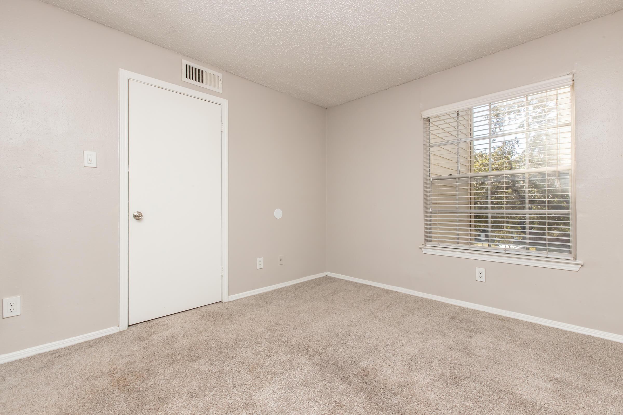 Empty interior of a light-colored room with beige carpet, featuring a closed white door, a window with blinds, and a neutral wall color. No furniture or decorations are present, creating a spacious and clean aesthetic.