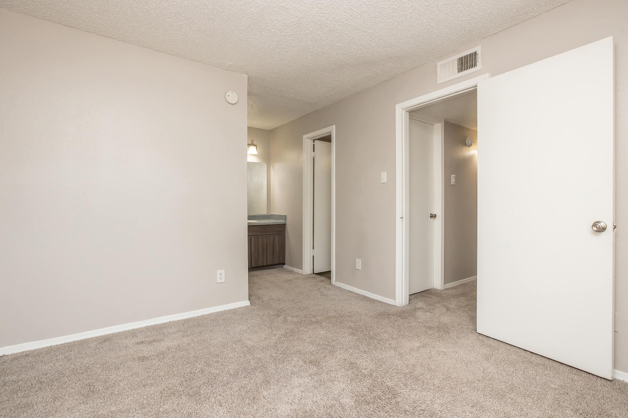 A spacious, neutral-colored room featuring beige carpet and light gray walls. There are two doorways leading to other rooms, and a bathroom area visible in the background with a sink and mirror. The room is well-lit with natural light.