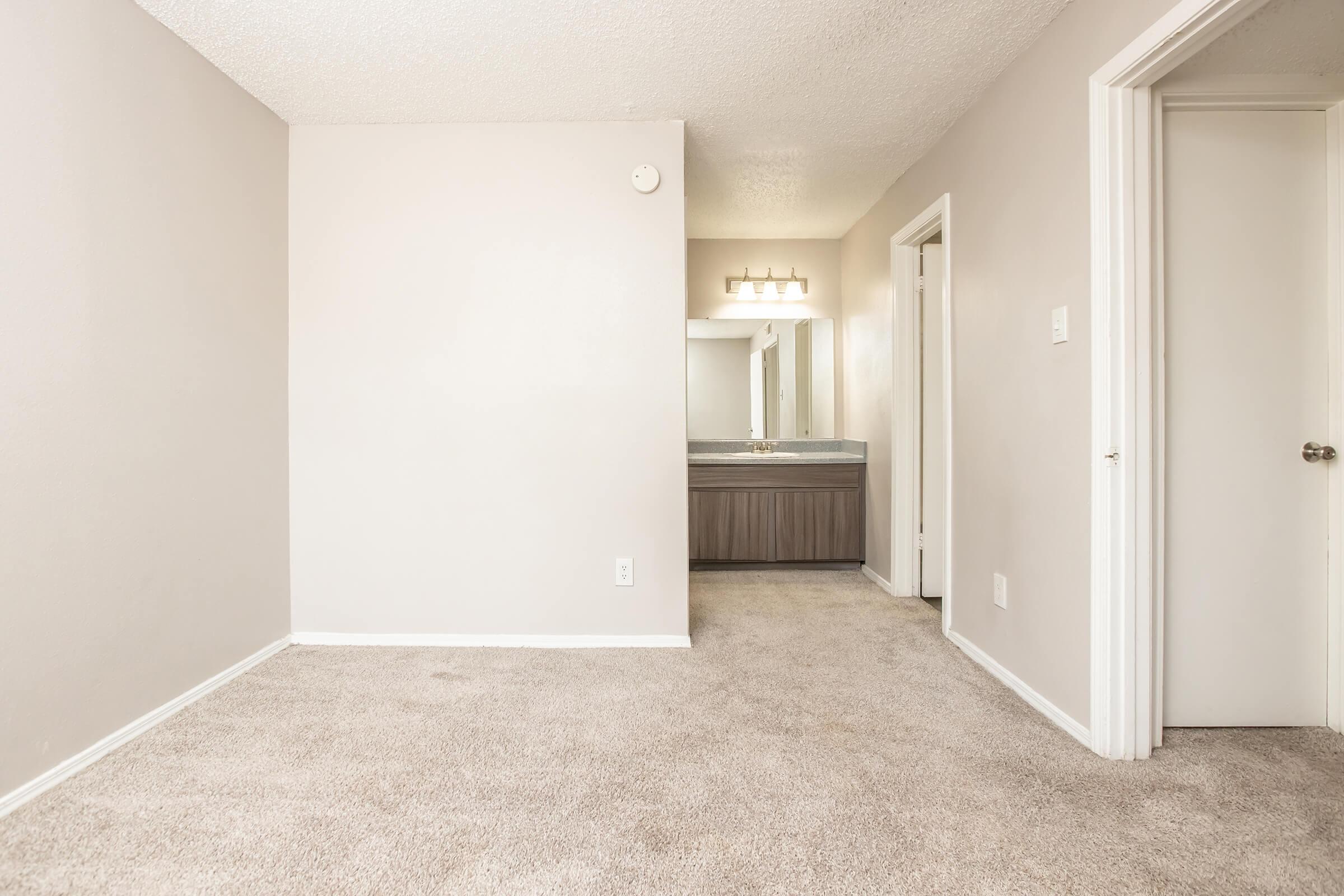 A spacious, empty room featuring light gray walls and plush beige carpet. In the background, there's a bathroom area with a large mirror and vanity. The room has two doorways leading to adjacent spaces, providing a sense of openness and potential for decoration or furniture arrangement.