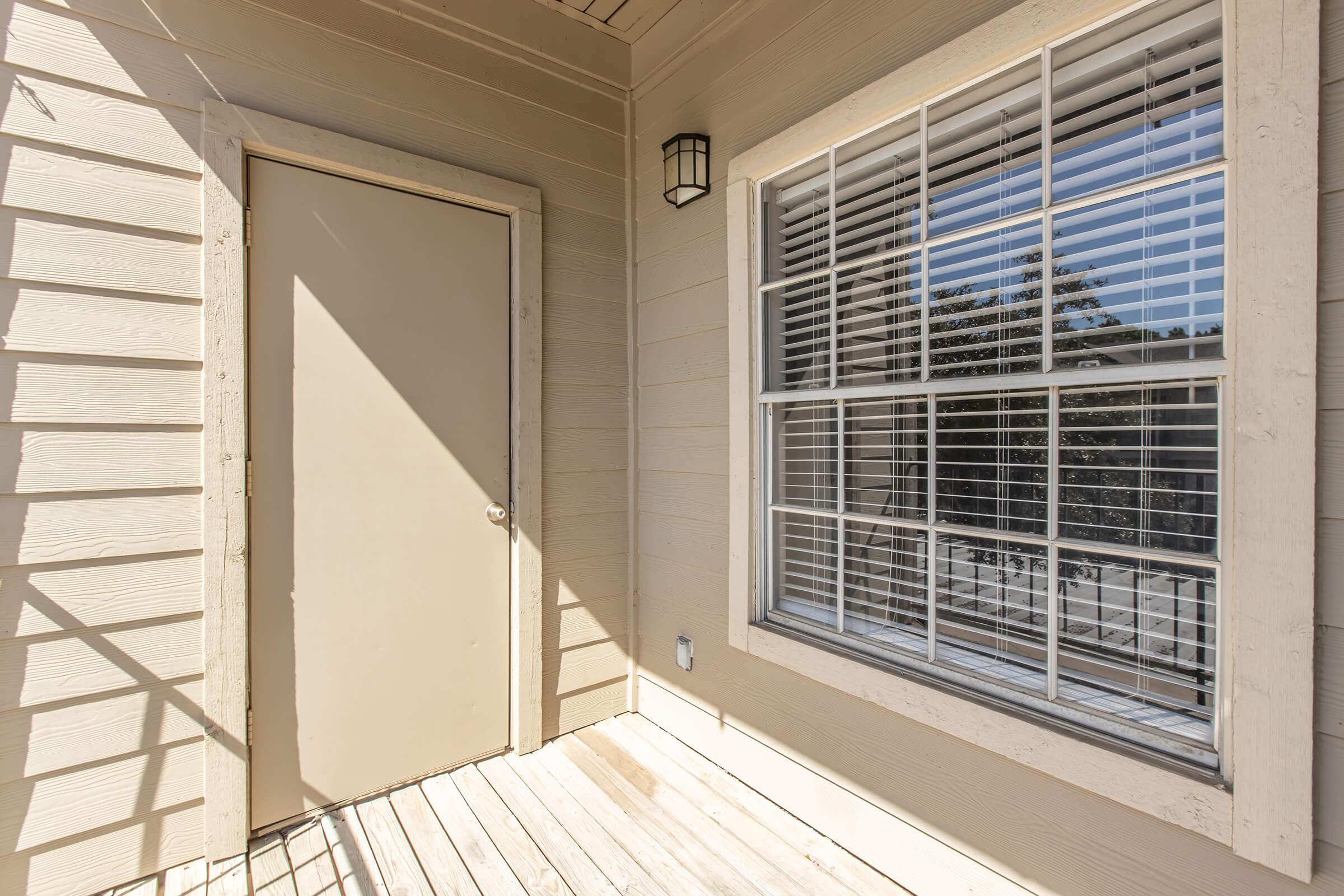 A small porch area featuring a sealed beige door and a window with white blinds. Sunlight casts shadows on the wooden floor, highlighting the textured wall and the outside greenery visible through the window.