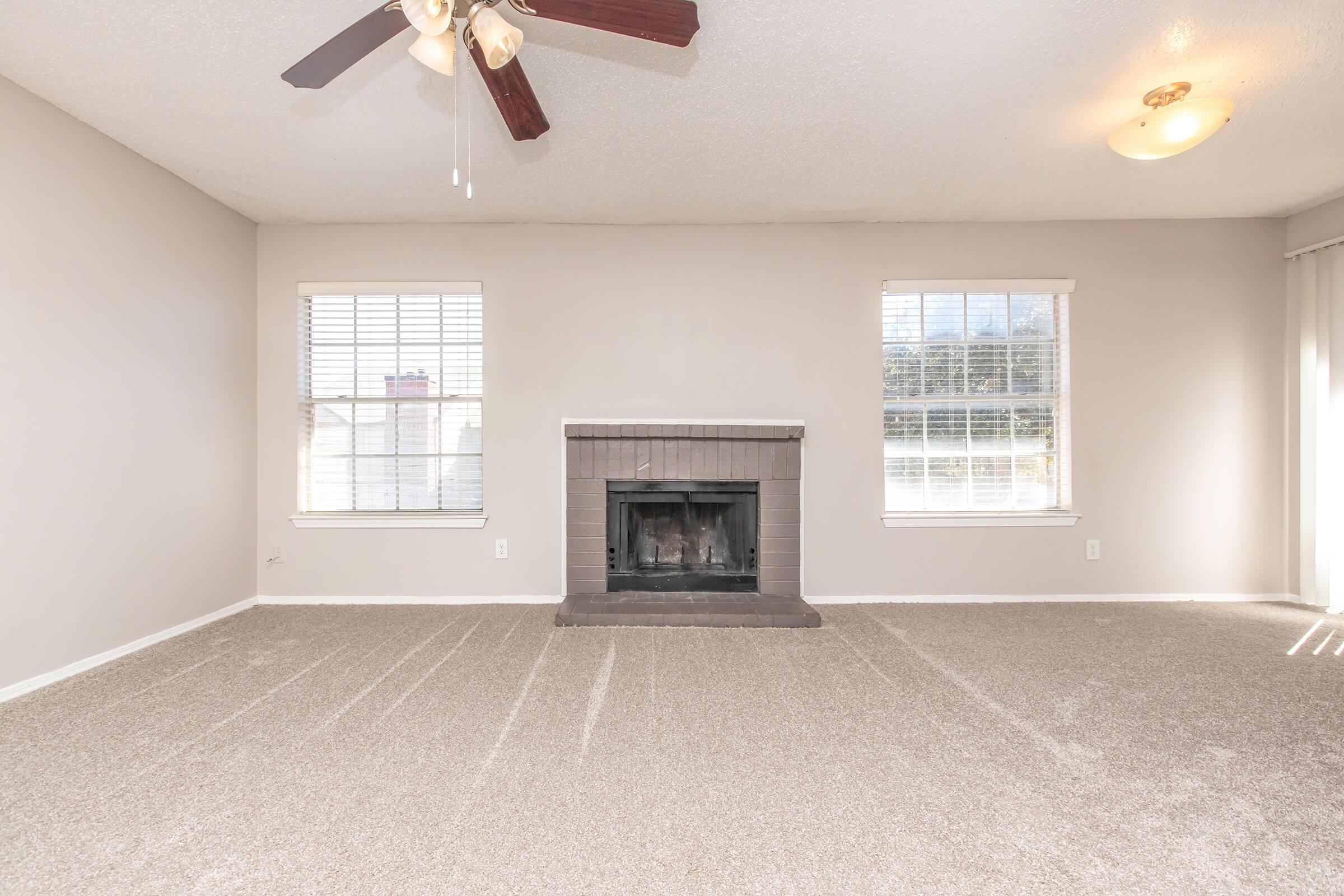 Empty living room with light-colored walls and beige carpet. Two windows provide natural light, and a ceiling fan is mounted above. A brick fireplace is centered on the back wall, with no furniture present, creating a spacious and airy atmosphere.