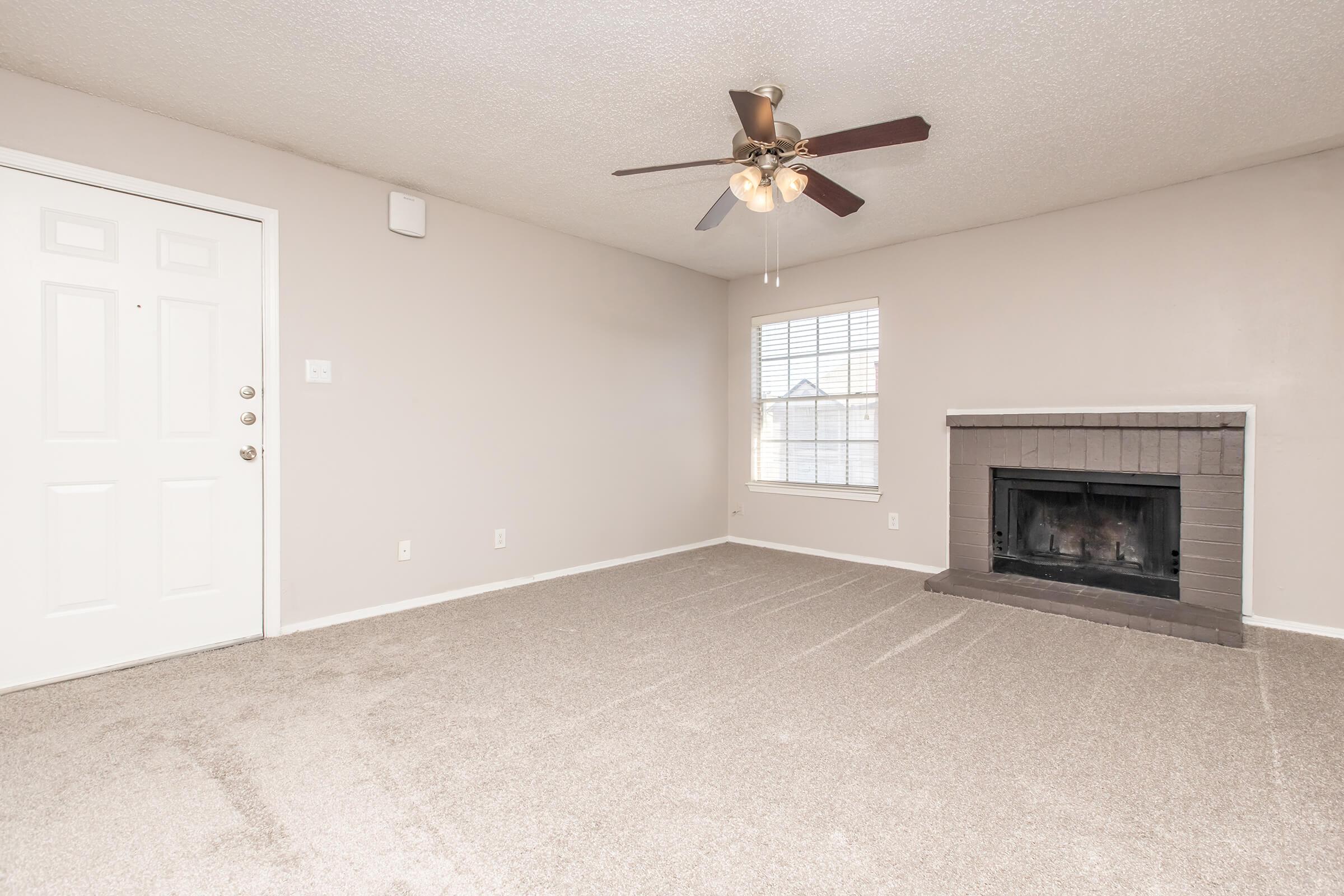 A spacious living room featuring light gray walls, a ceiling fan with lights, a window with blinds, and a cozy fireplace. The floor is covered with beige carpet, and there's an entry door on the left side.