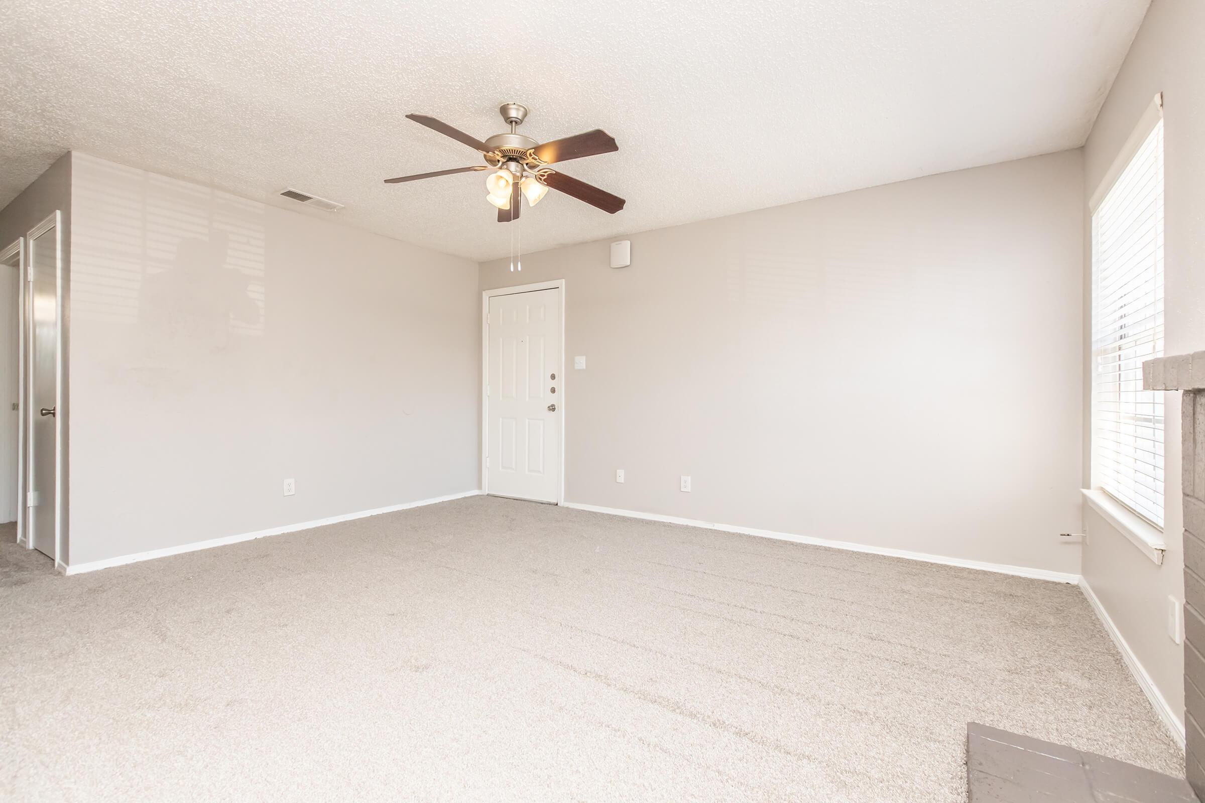 A vacant room with light gray walls and carpet, featuring a ceiling fan with wooden blades. There is a door on the left, a white front door in the center, and a window on the right allowing natural light. The space appears clean and ready for furnishing.
