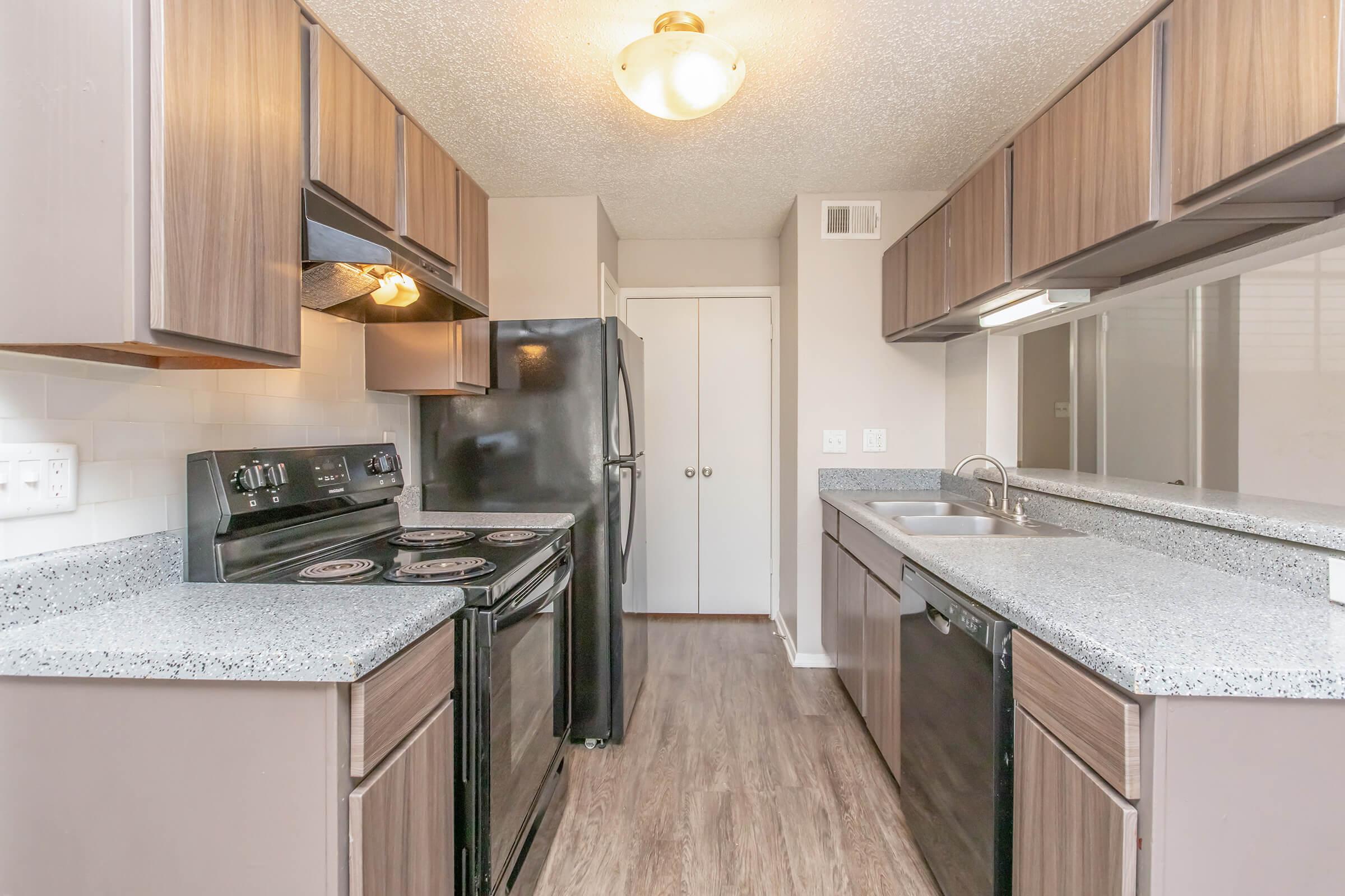 Modern kitchen featuring brown wood cabinetry, black appliances including a stove and refrigerator, and a gray speckled countertop. The space is well-lit with a ceiling fixture, and a sink is visible next to the dishwasher. The floor has a wood-like finish, creating a warm and inviting atmosphere.