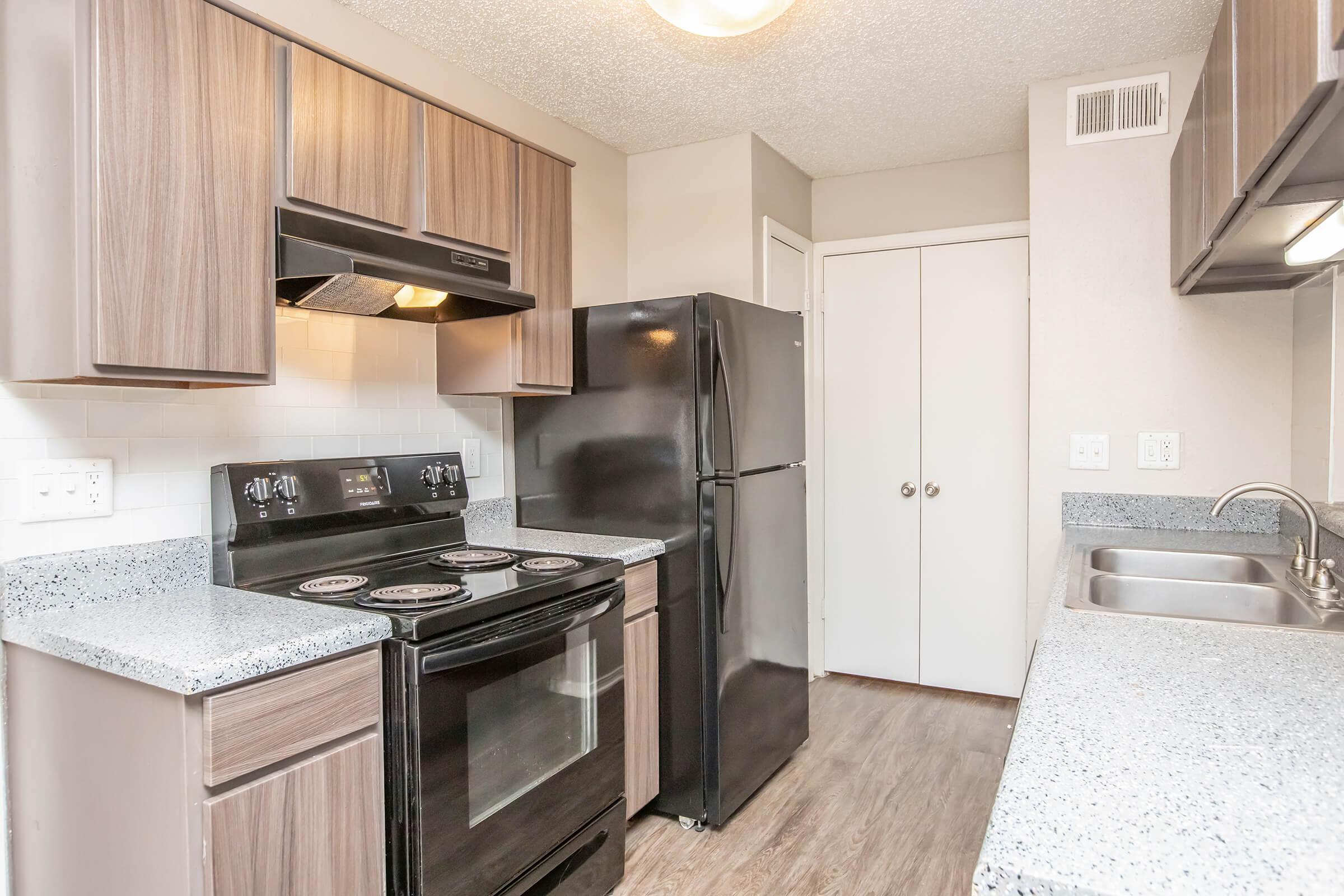A modern kitchen featuring wooden cabinets, a black refrigerator, a black stove with four burners, and a stainless steel sink. The countertops are made of speckled gray material, and the walls are painted in neutral tones. The space is well-lit with overhead lighting.