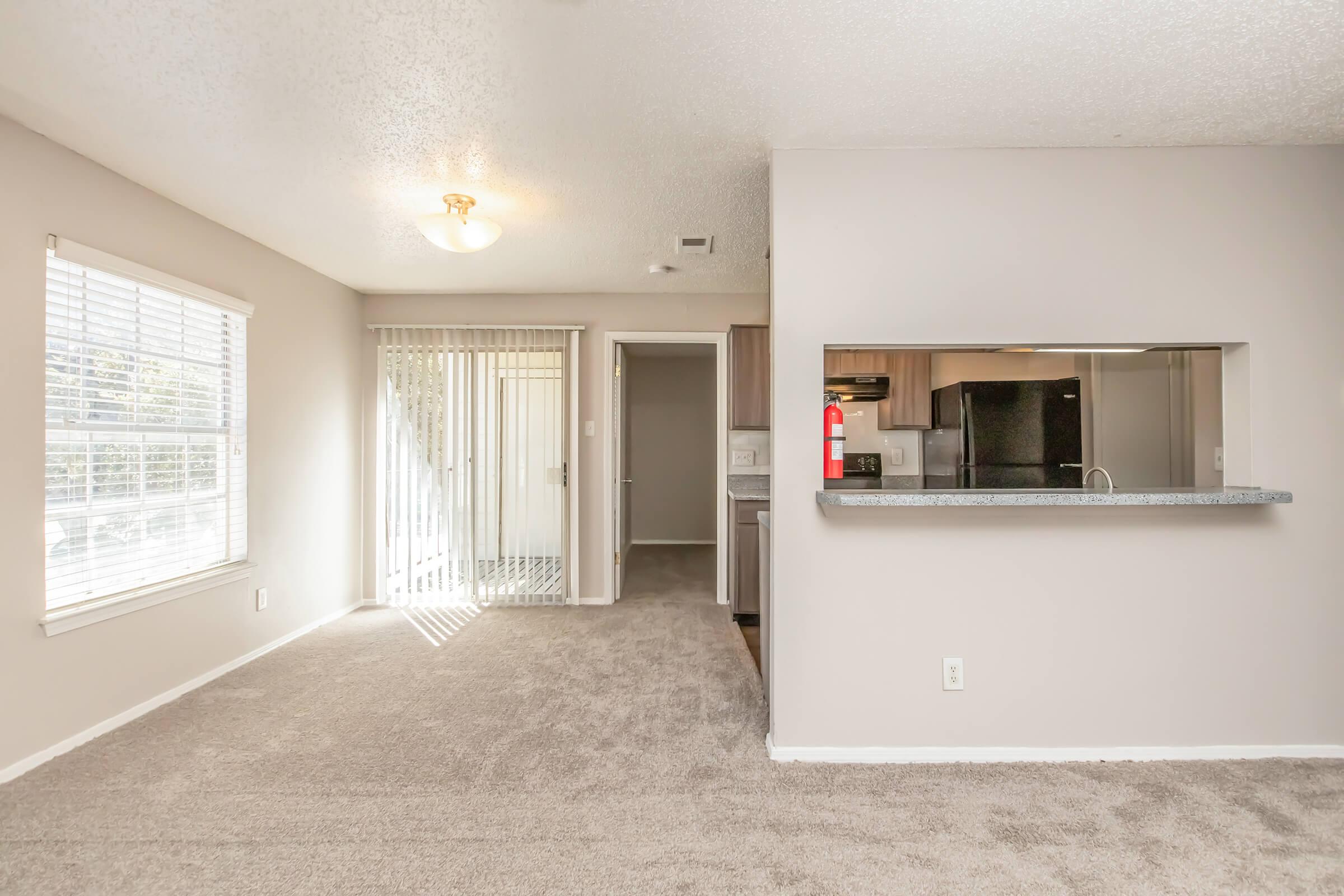 A spacious living area with light gray carpet, a large window allowing natural light, and a sliding glass door leading to an outdoor space. The kitchen is partially visible, featuring modern appliances. The walls are painted in a neutral tone, creating a cozy atmosphere.