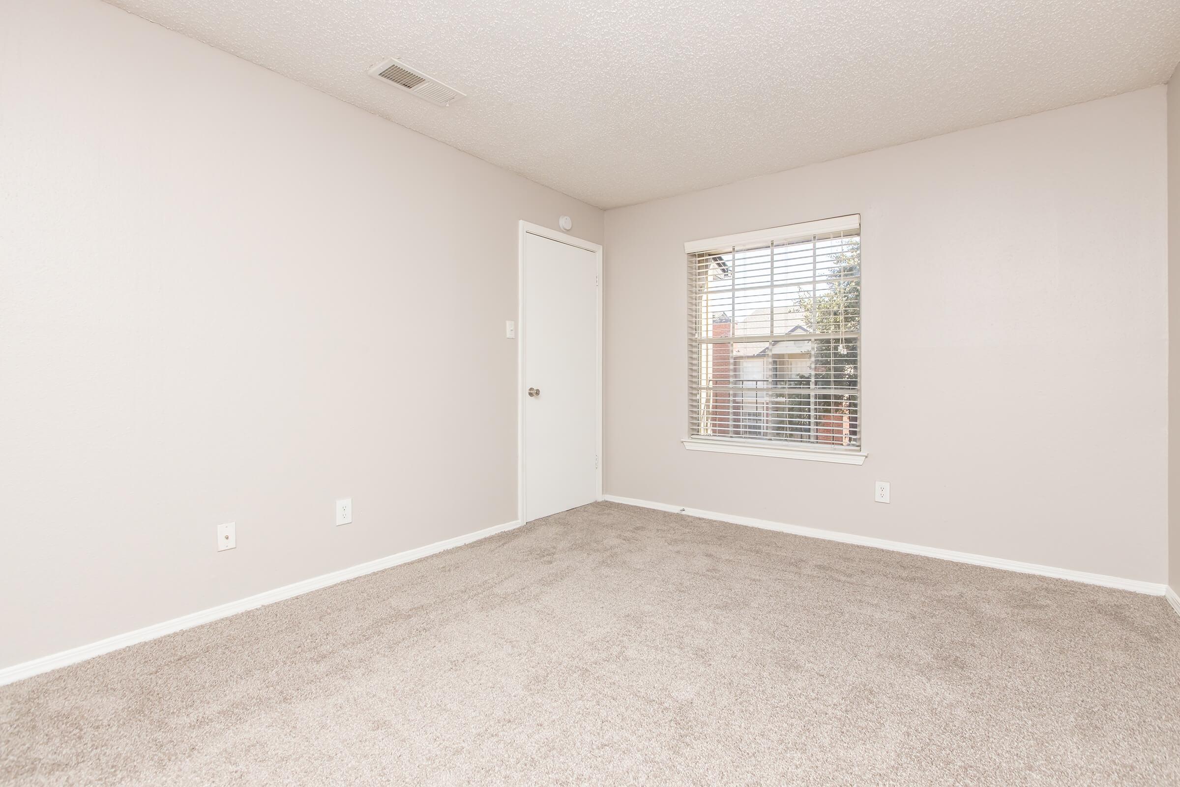 Empty room with light gray walls and carpet. There is a closed door on the left and a window with blinds on the right, showing trees outside. The room has no furniture and features a simple, clean aesthetic.