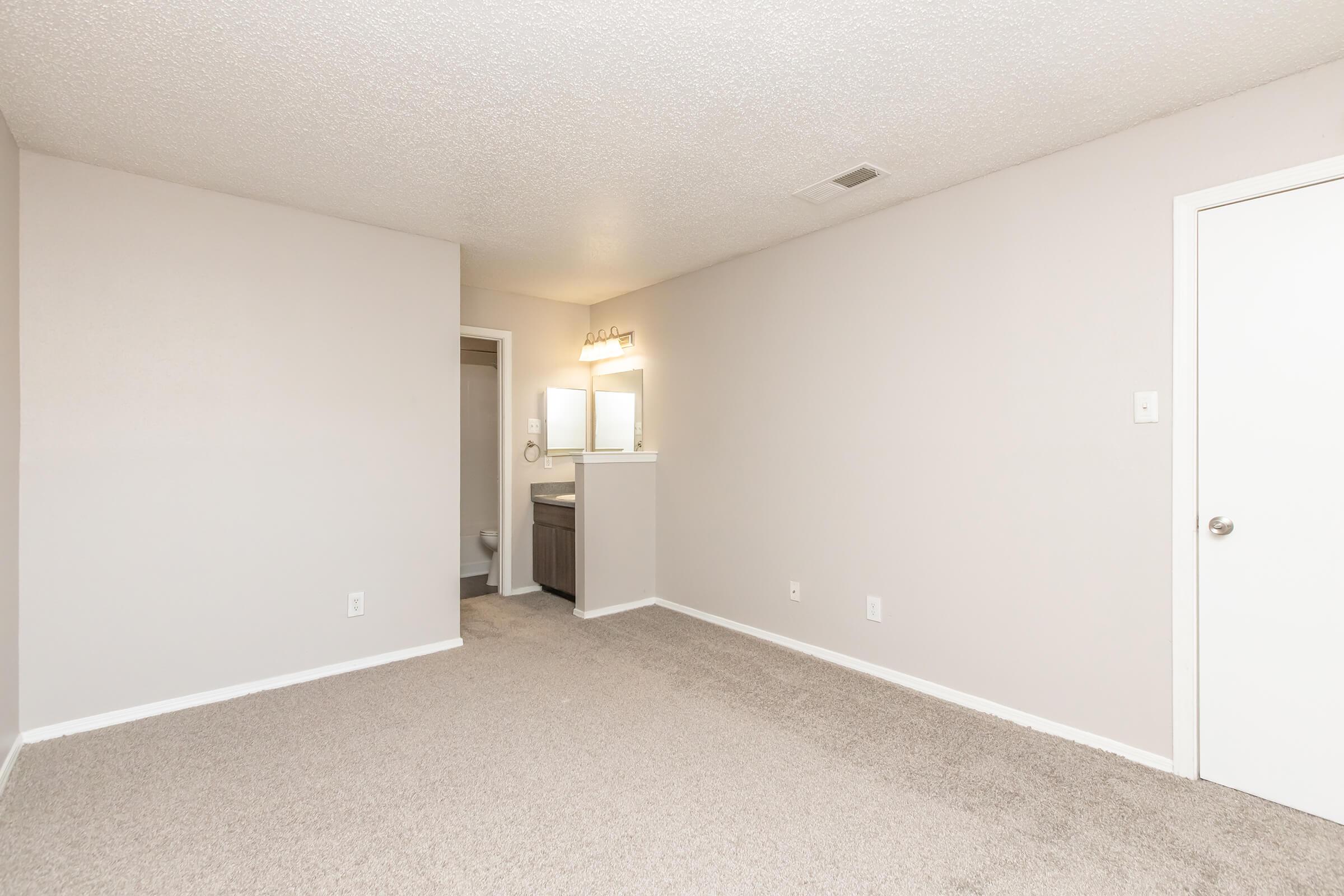 A well-lit, empty room with light gray walls and carpet flooring. To the left, there is a small alcove with a bathroom visible. The bathroom features a mirror and sink area. The room has no furniture, creating a spacious, open feel.
