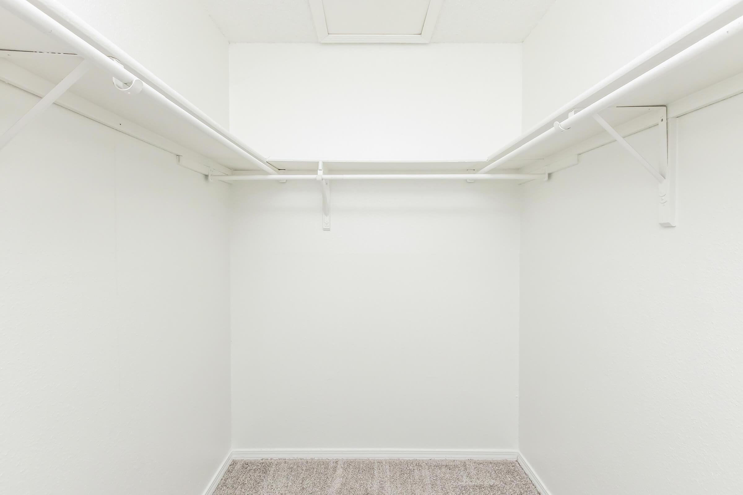 Empty walk-in closet with white walls and a carpeted floor. Two white shelves are mounted on the walls, providing storage options. The space is well-lit and has a clean, minimalist appearance.