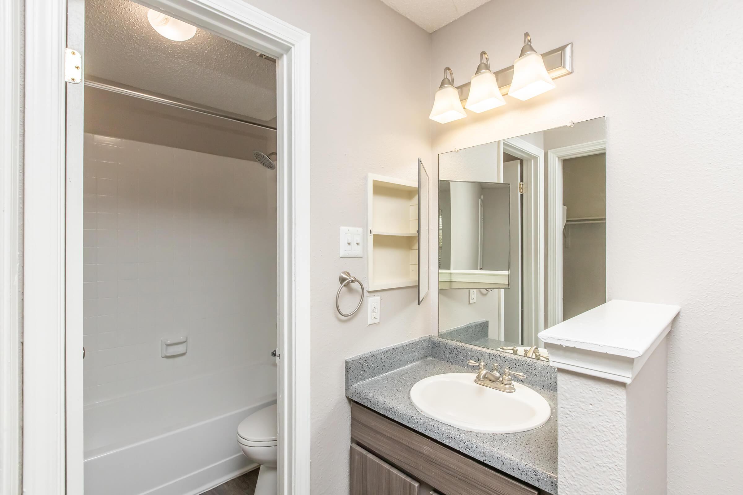 A view of a modern bathroom featuring a white bathtub with a shower curtain, a single sink with a granite countertop, a wall-mounted mirror, and a light fixture. The walls are painted in a light color, and there is a towel ring near the sink. A door leads to a closet space.