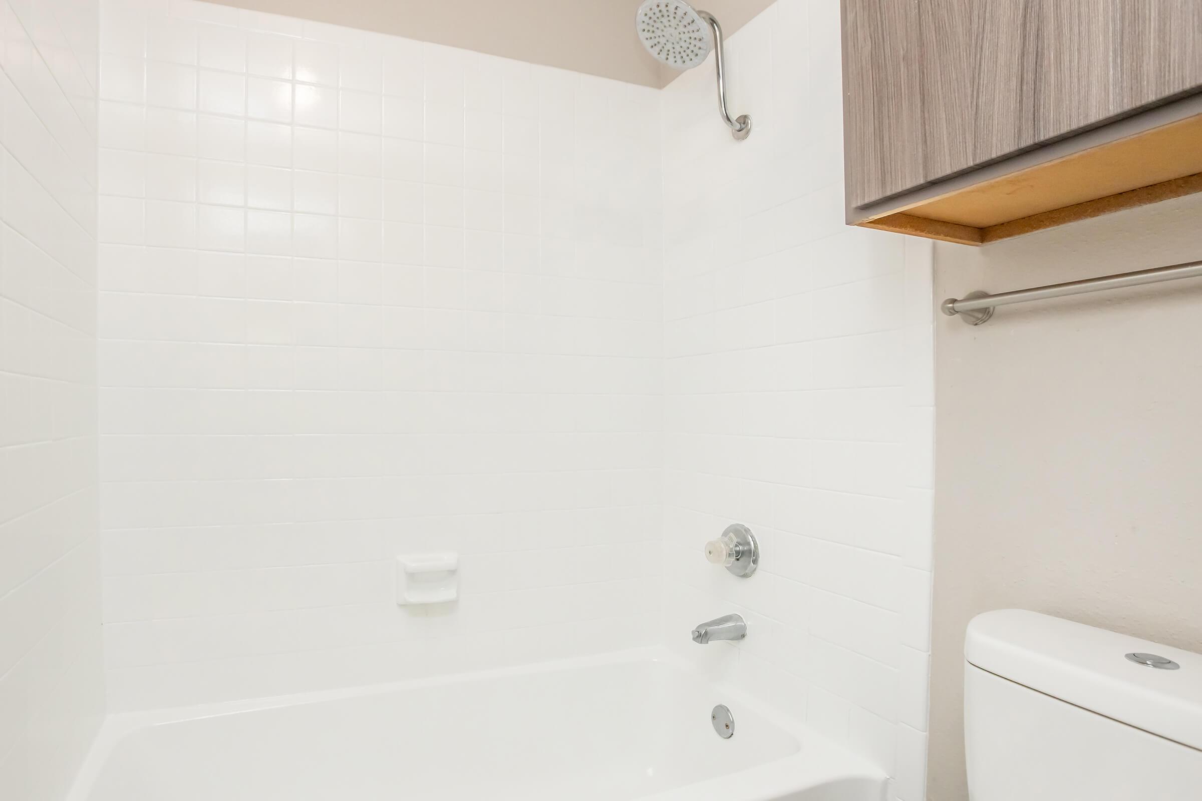 A clean, minimalist bathroom featuring a white tiled shower with a showerhead, a white bathtub, a modern toilet, and a wooden cabinet above. The space is bright and uncluttered, emphasizing a contemporary design.