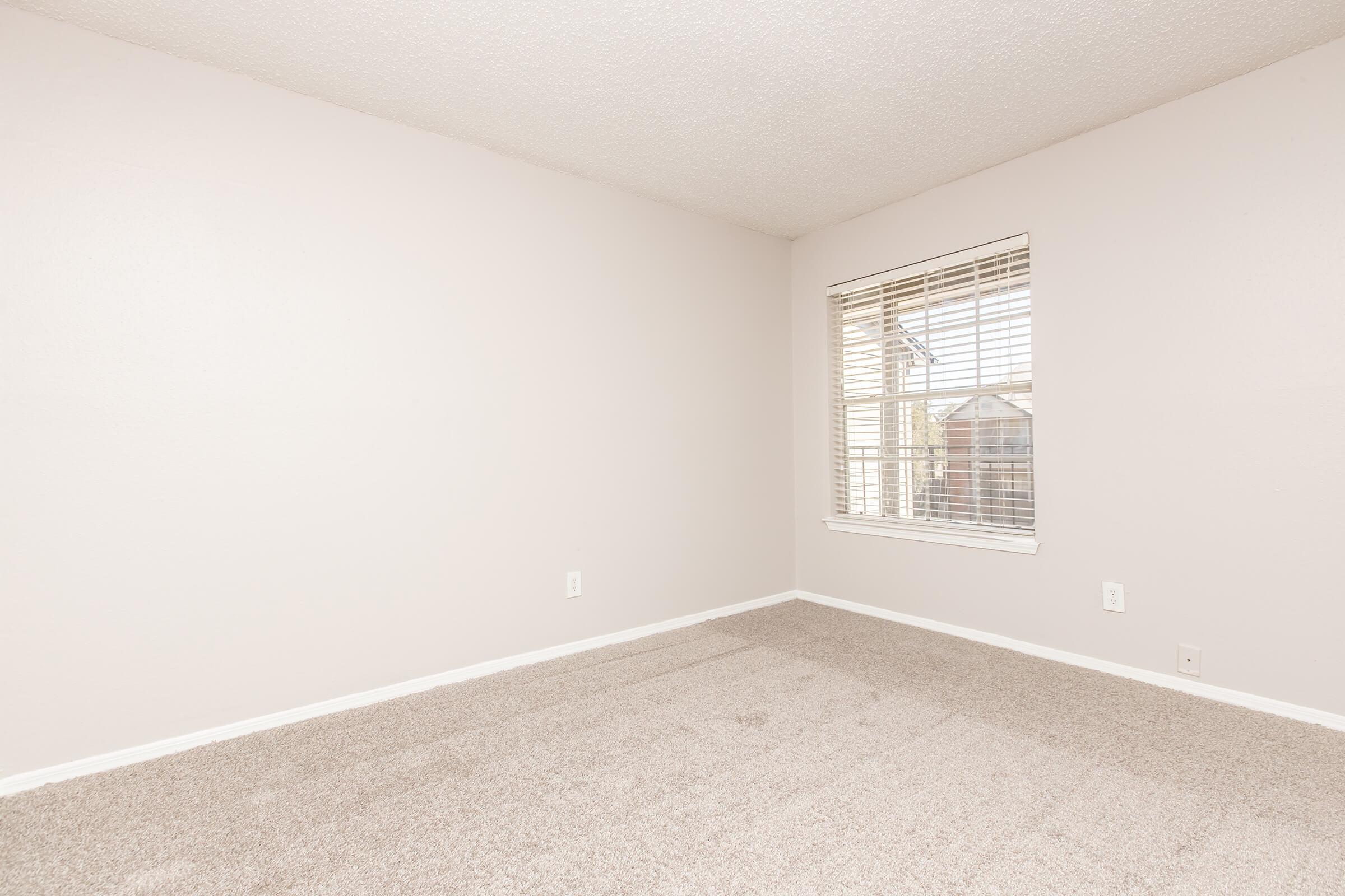 An empty, well-lit room with light beige walls and a window featuring blinds. The floor is covered with soft, neutral-colored carpet, and there are no furniture or decorations visible. The room appears spacious and inviting, with natural light coming in from the window.
