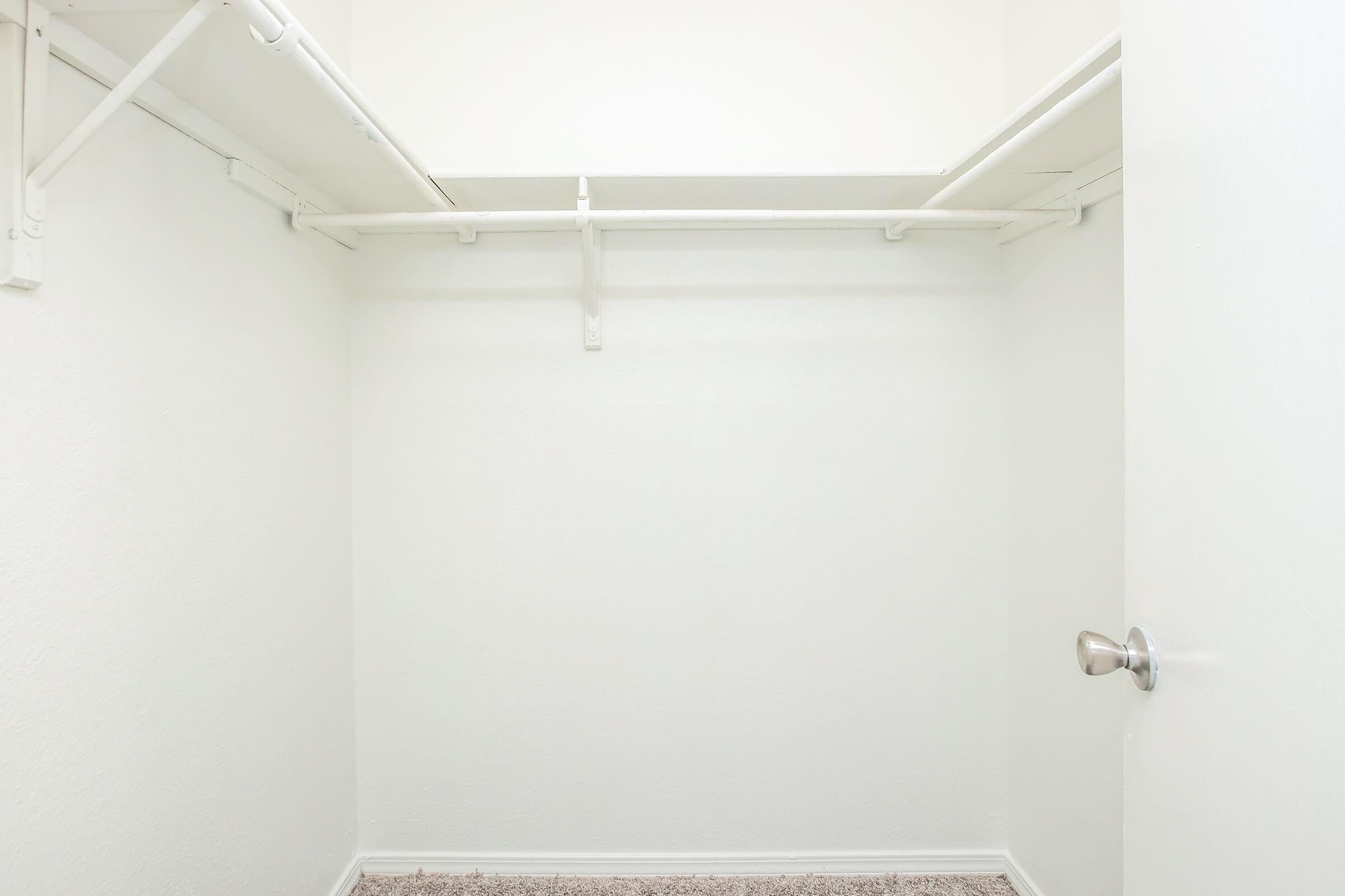 Empty closet with white walls and a light carpeted floor. There are metal shelving units along the top, and a closed door is visible on the right side. The overall appearance is neat and minimalistic.