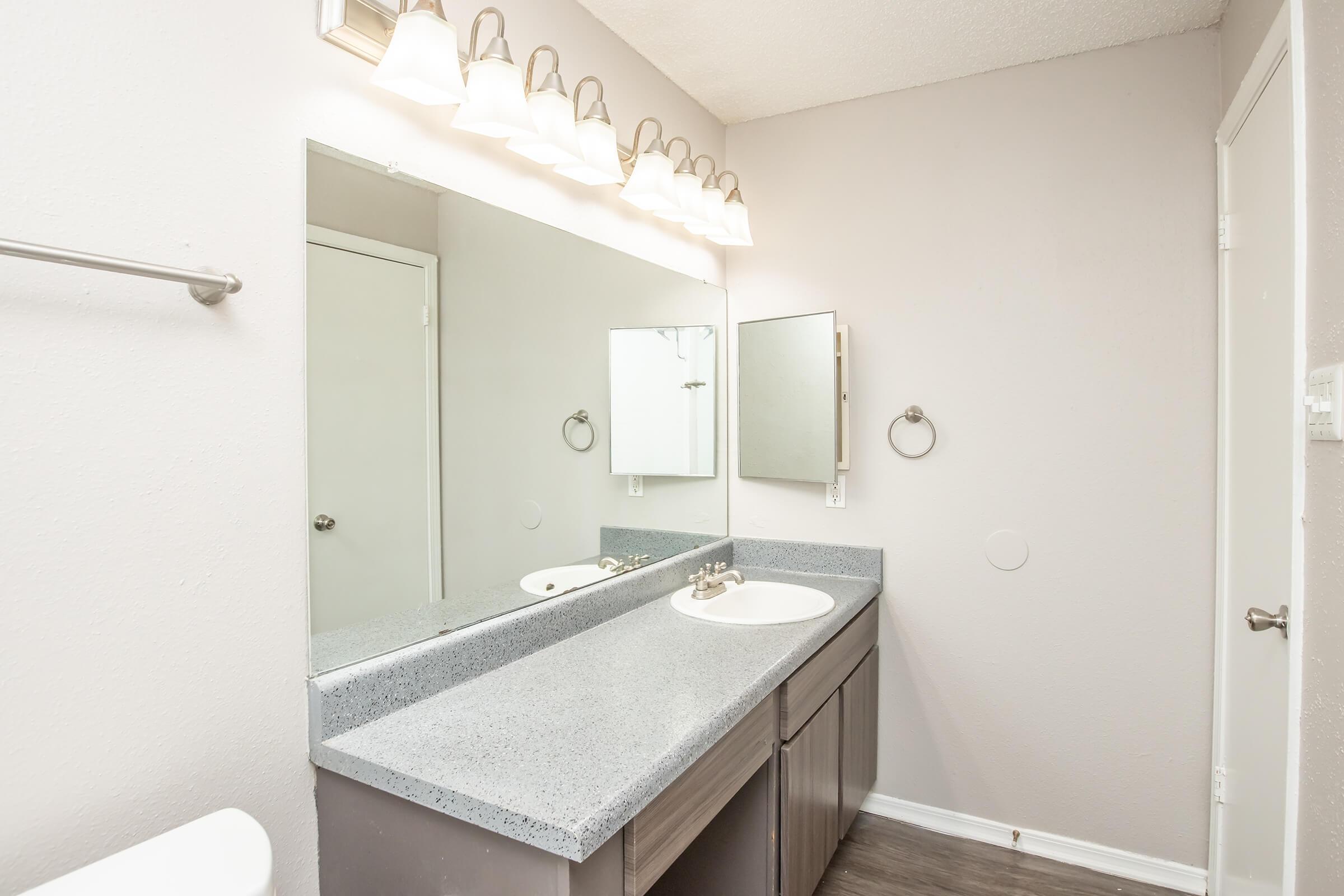 A modern bathroom featuring a double sink vanity with gray cabinets and a light granite countertop. Above the sinks, there are five light fixtures installed on the wall. A large mirror spans the wall above the sinks, while the walls are painted in a soft gray color, and the floor is a dark wood laminate.
