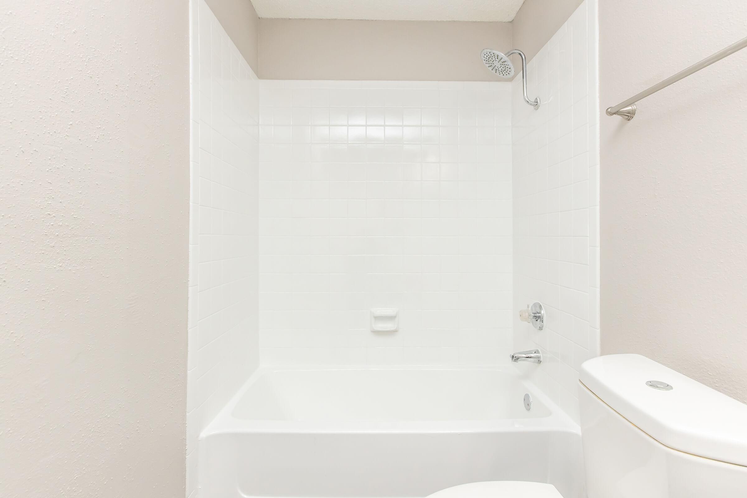A clean, minimalist bathroom featuring a white tiled shower and bathtub combo, a modern chrome showerhead, and a white toilet. The walls are painted in a soft neutral color, creating a bright and open space. No additional decor or accessories are present.