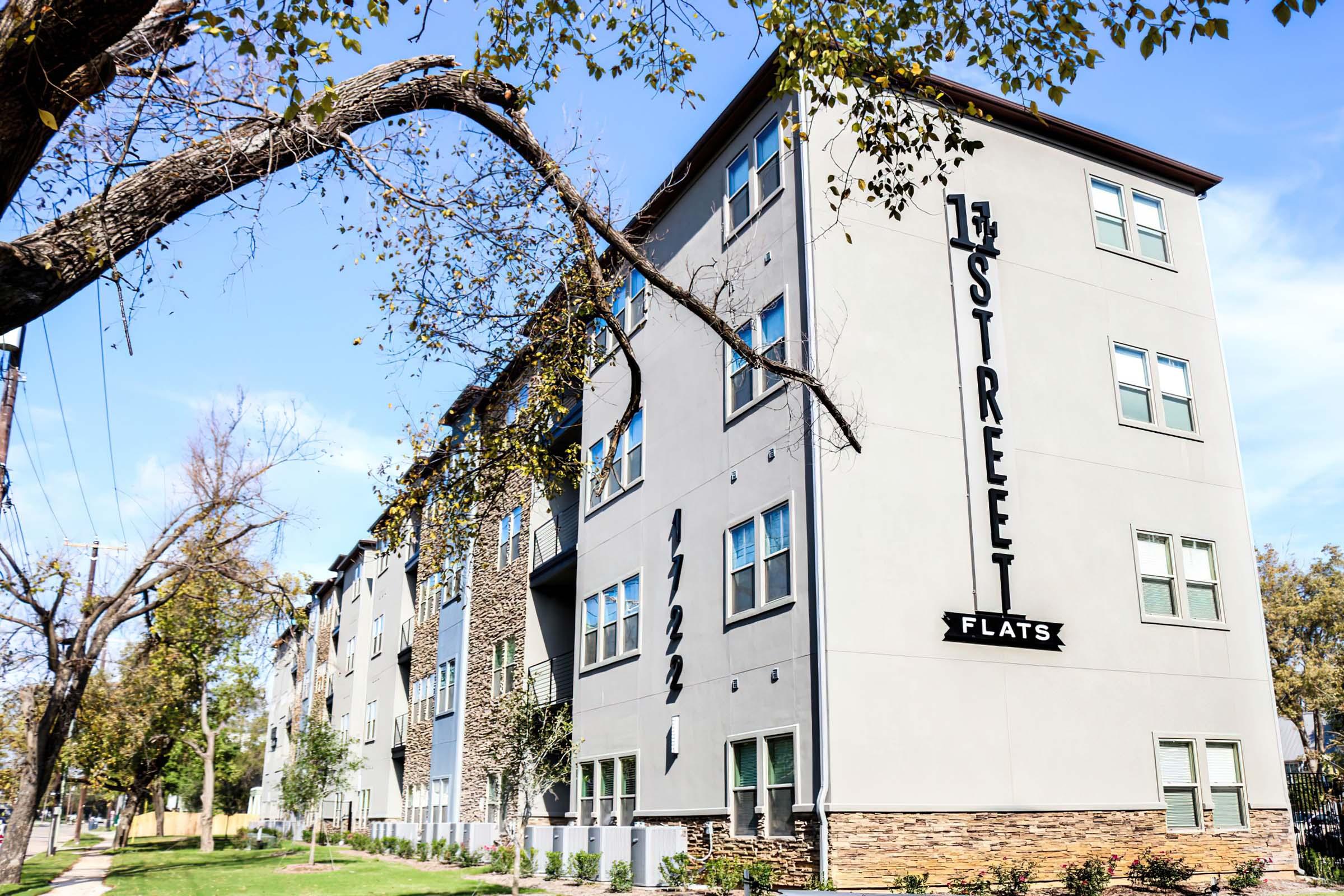 Modern apartment building labeled "1st Street Flats," featuring large windows and a mix of stone and stucco exteriors. The structure has three levels and is surrounded by trees and landscaped grass areas under a clear blue sky.