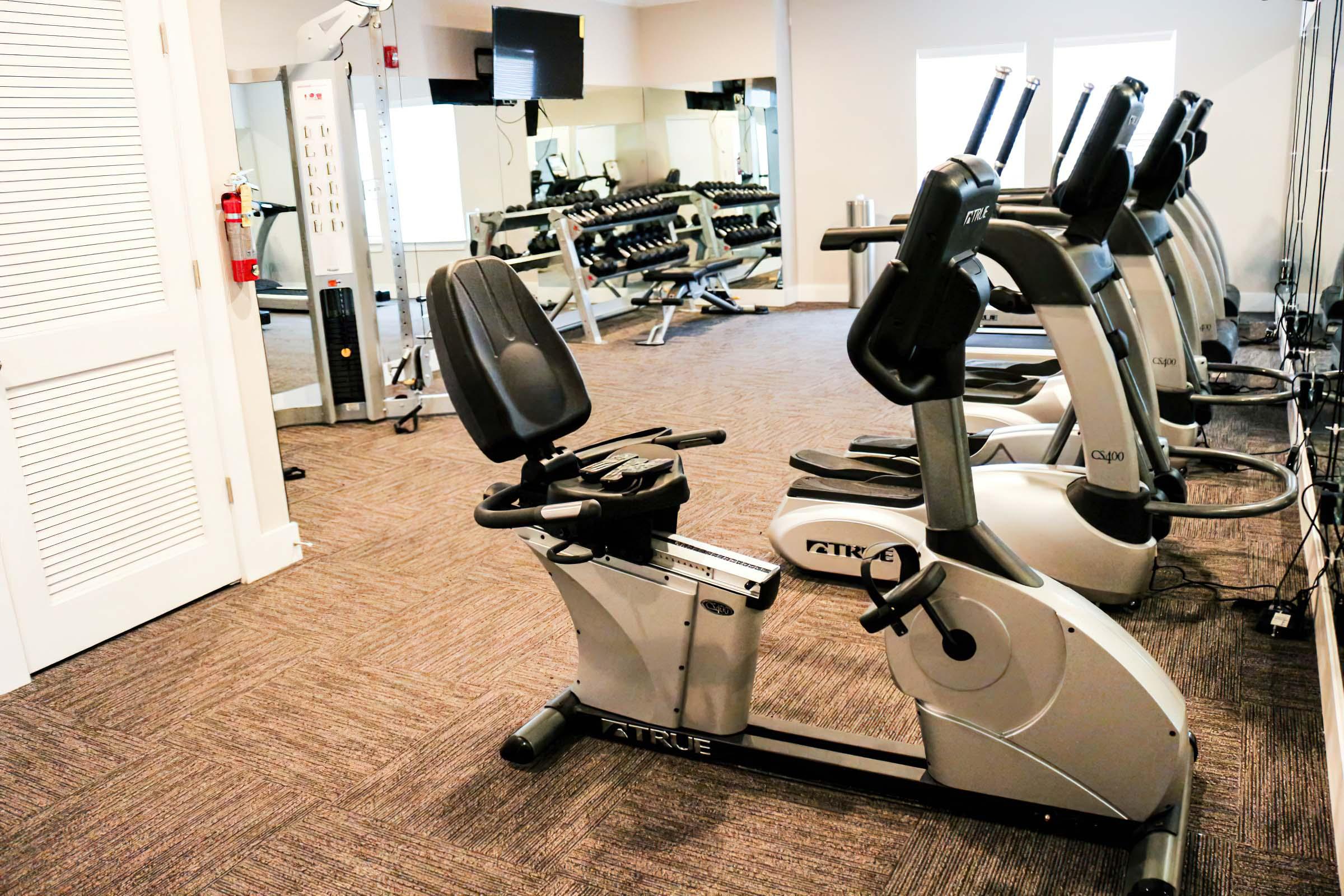 A fitness area featuring several exercise machines, including a recumbent bike in the foreground. The background includes a set of dumbbells on a rack and mirrors, creating a well-lit workout space with carpet flooring.