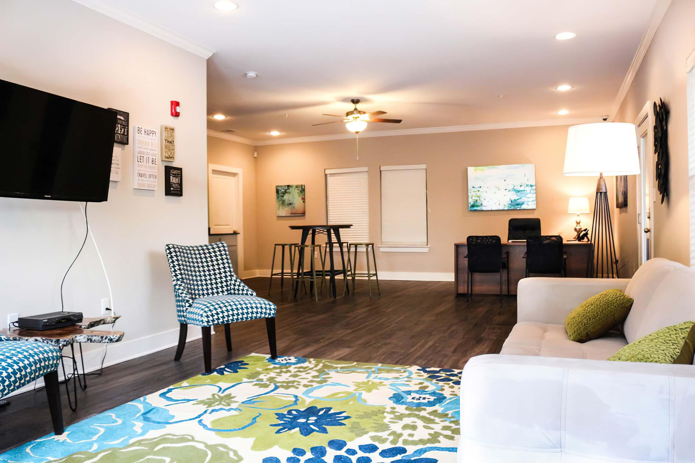 A modern living room featuring a comfortable couch, two patterned chairs, and a coffee table. A large flat-screen TV is mounted on the wall. The floor is covered with a colorful area rug, and there are decorative wall art pieces. Natural light comes in through windows, creating a bright, inviting atmosphere.