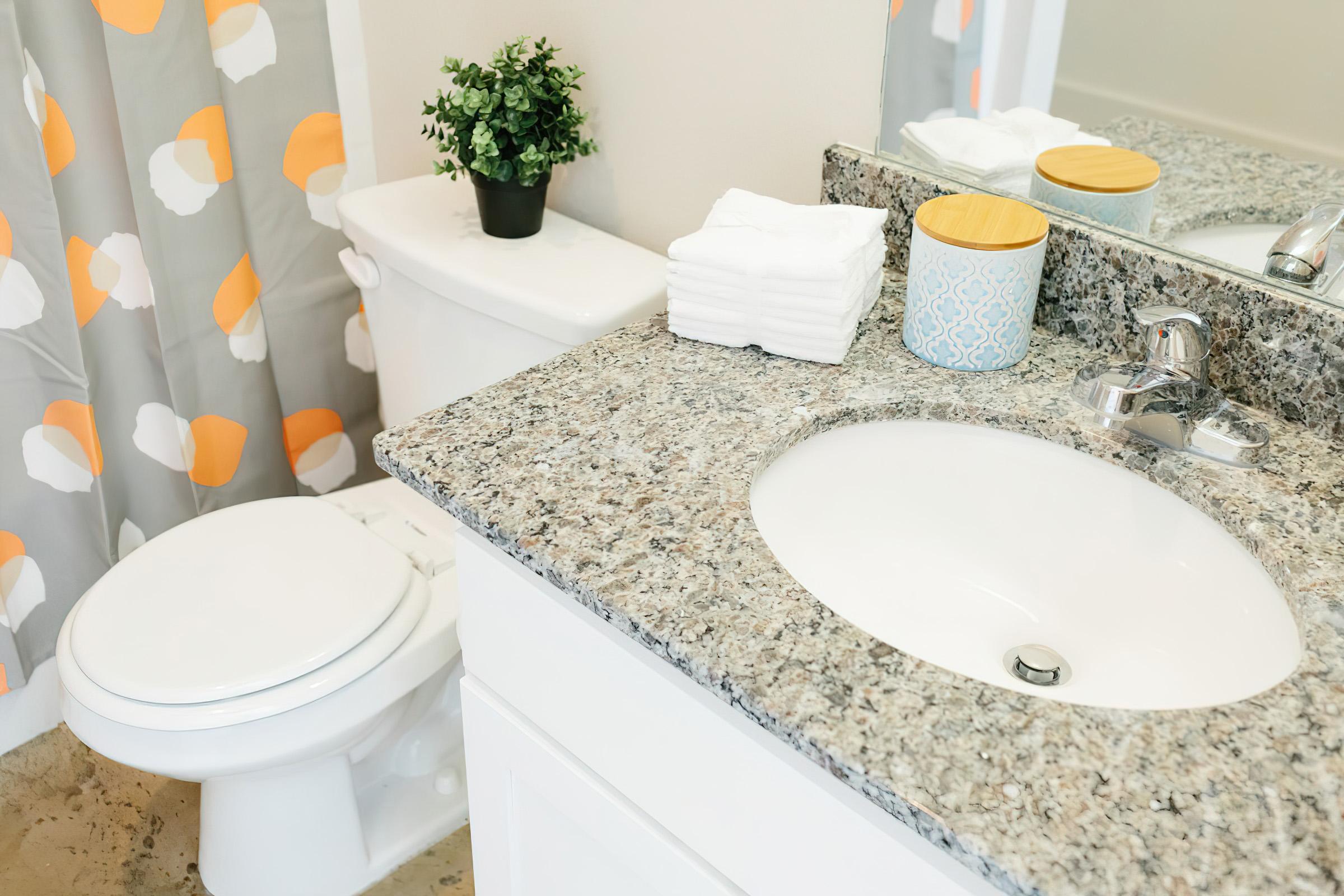 A clean bathroom featuring a white toilet, a granite countertop with a bowl sink, neatly stacked white towels, a small potted plant, and a decorative container. A shower curtain with a colorful pattern adds a bright touch to the space.