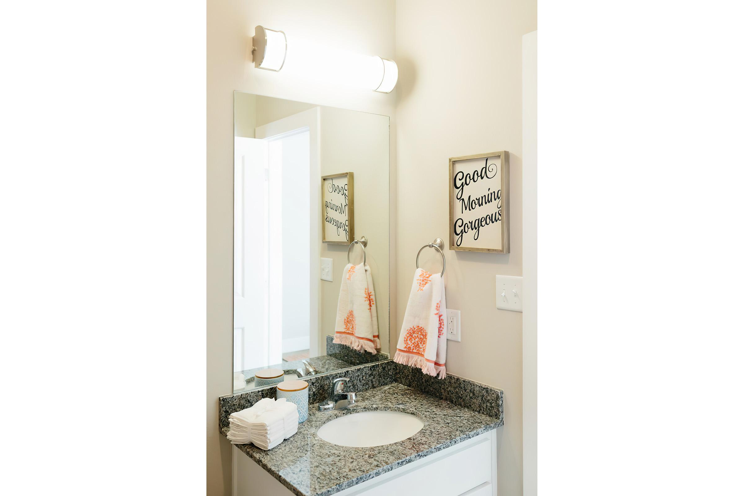 A modern bathroom featuring a granite countertop with a sink, a large mirror, and a light fixture above. Decorative towels hang on a towel rack, and a framed sign reading "Good Morning Gorgeous" is displayed on the wall. Fresh white washcloths are neatly arranged on the countertop.