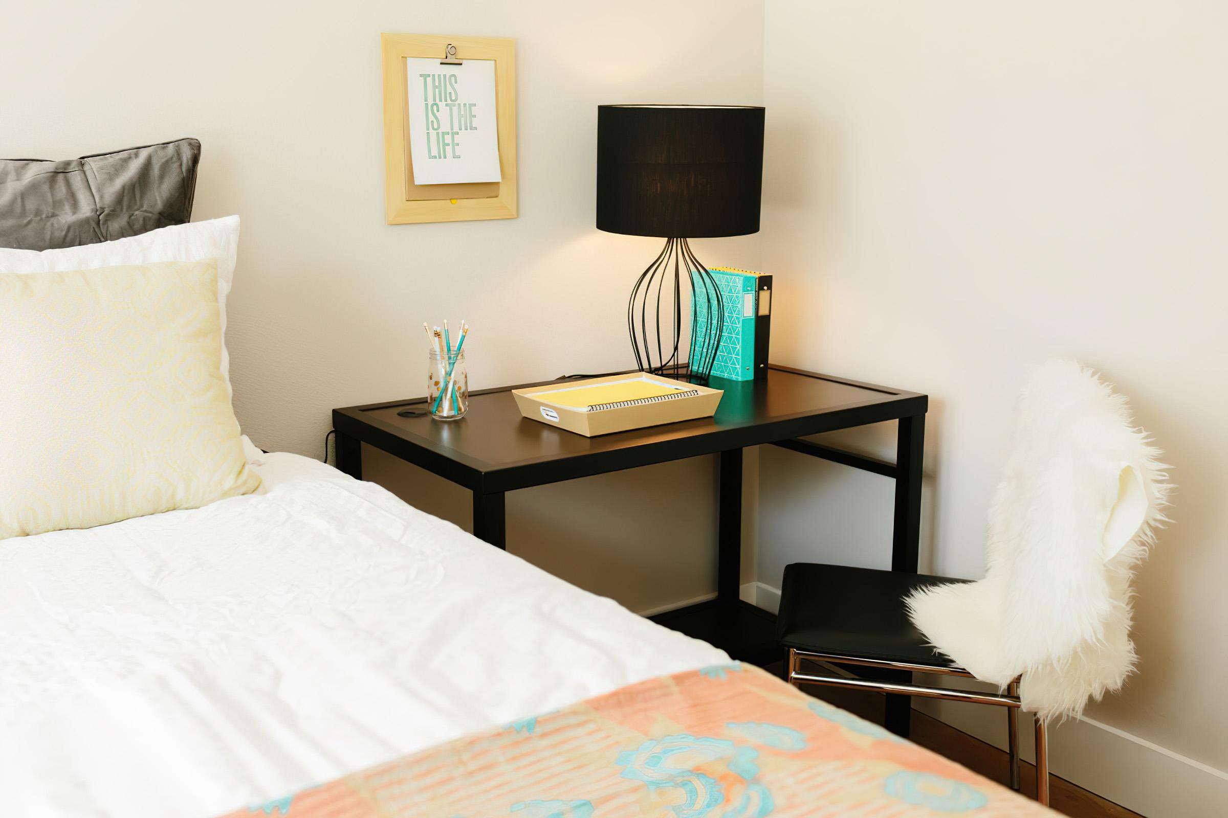 A cozy bedroom scene featuring a neatly made bed with white bedding, a small wooden desk with a lamp, and a colorful notebook. The desk is adorned with writing utensils and books, alongside a stylish chair with a fluffy white seat cover. A framed quote on the wall adds a personal touch to the space.